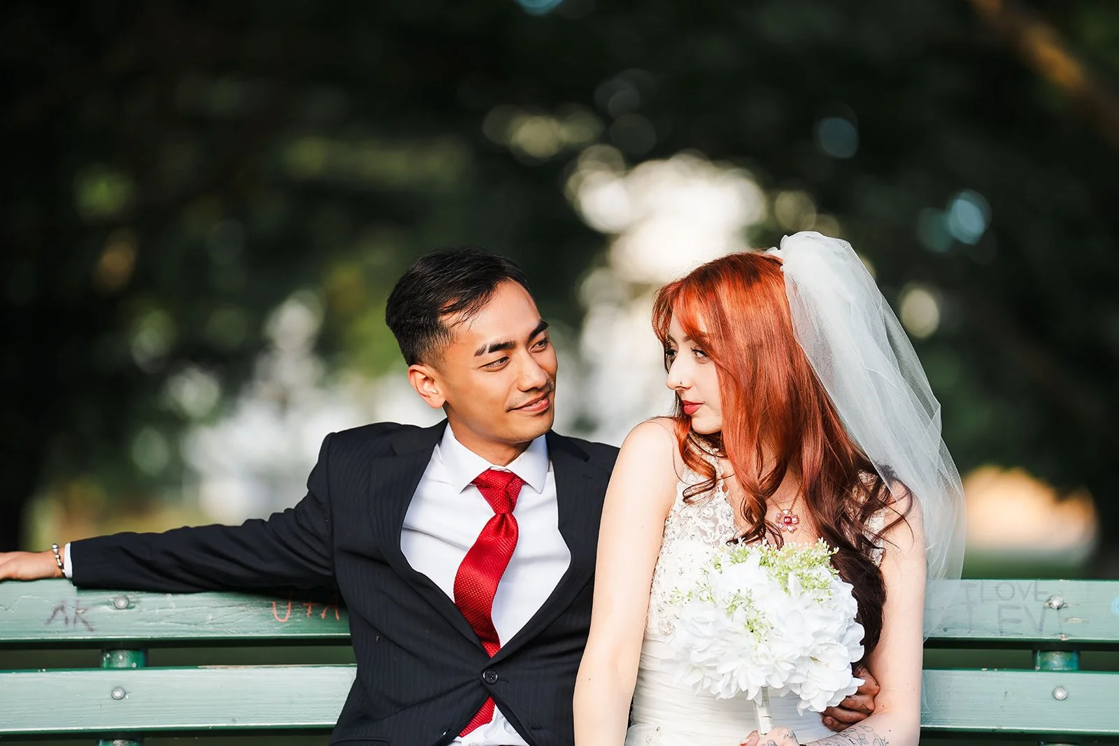 A wedding couple sitting on a park bench. The groom, wearing a black suit and red tie, looks at the bride. The bride, with long red hair and a white dress with a veil, holds a bouquet and gazes at the groom. Trees and blurred background in the park.