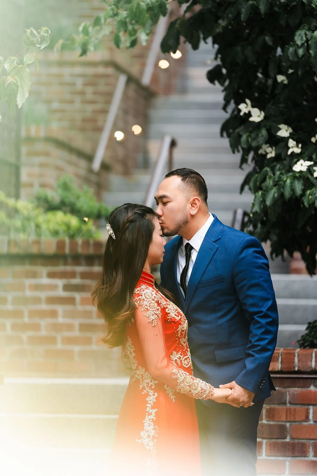 A couple holding hands; the man is kissing the woman's forehead outdoors near stairs and greenery.