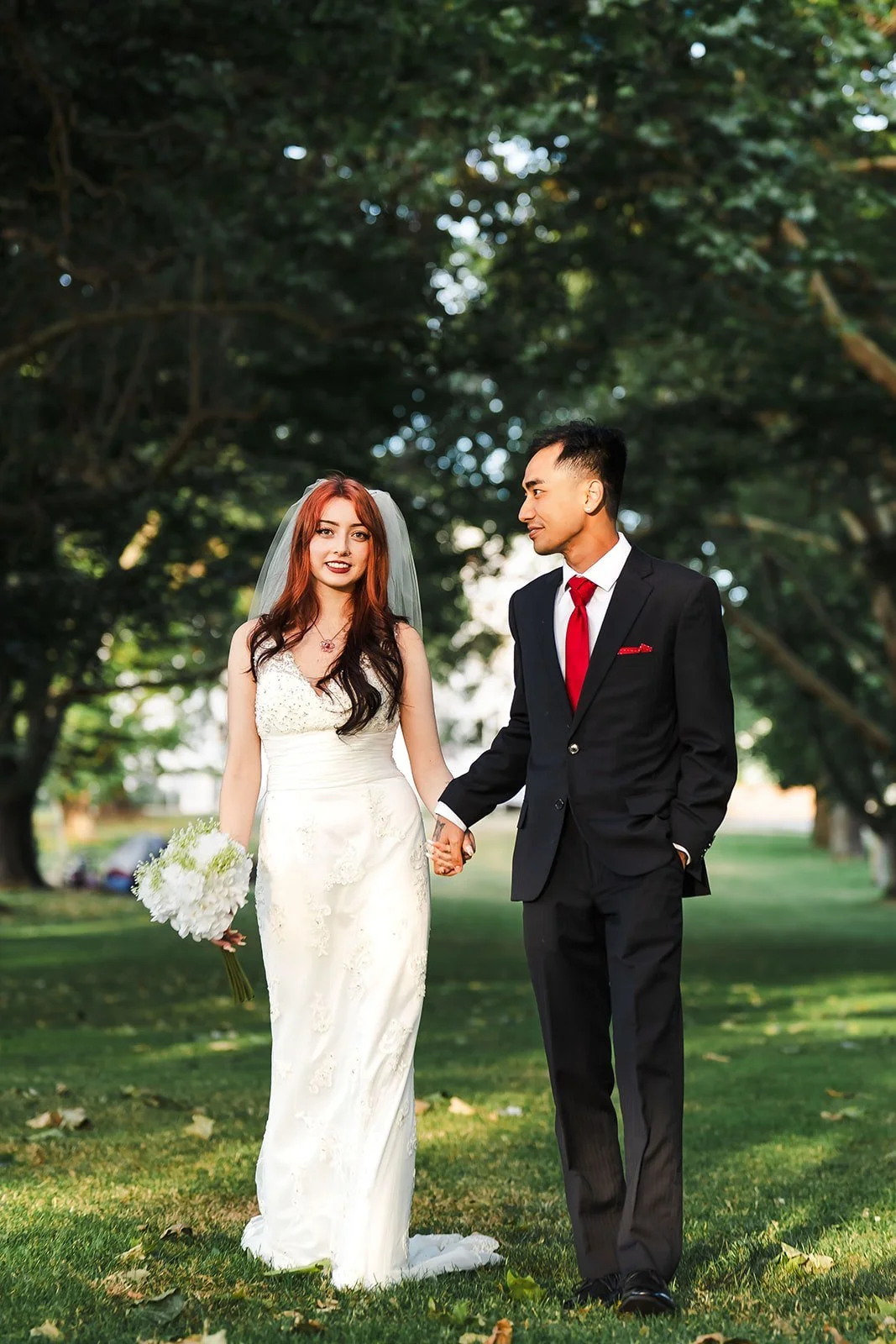 A bride and groom holding hands in a park, the bride in a white wedding gown holding a bouquet, and the groom in a black suit with a red tie.