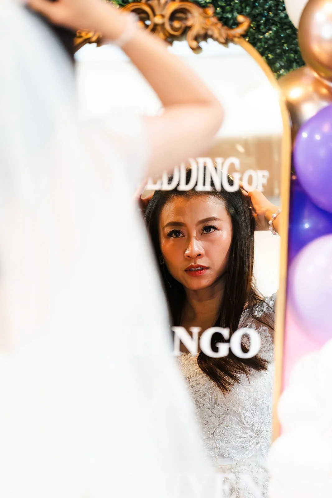 Woman in white dress looking at her reflection in a mirror with 'Wedding of' written on it, surrounded by colorful balloons.