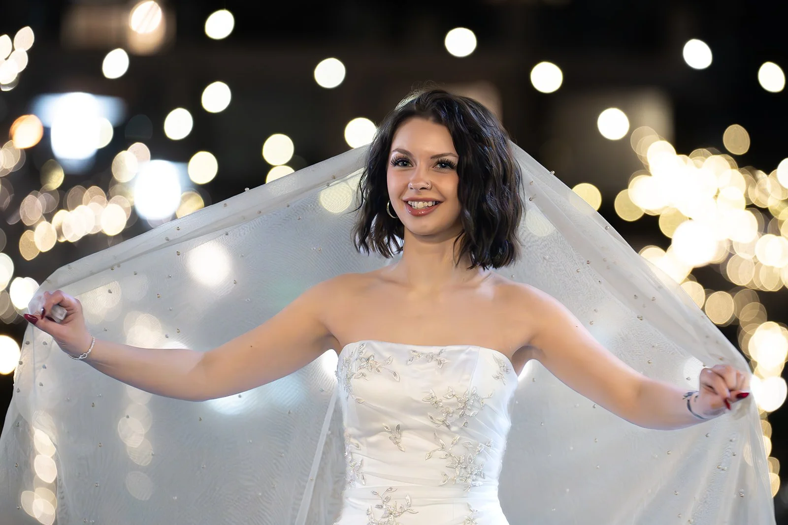 A smiling woman in a strapless white wedding dress with embroidery, holding her veil outstretched against a backdrop of blurred string lights.