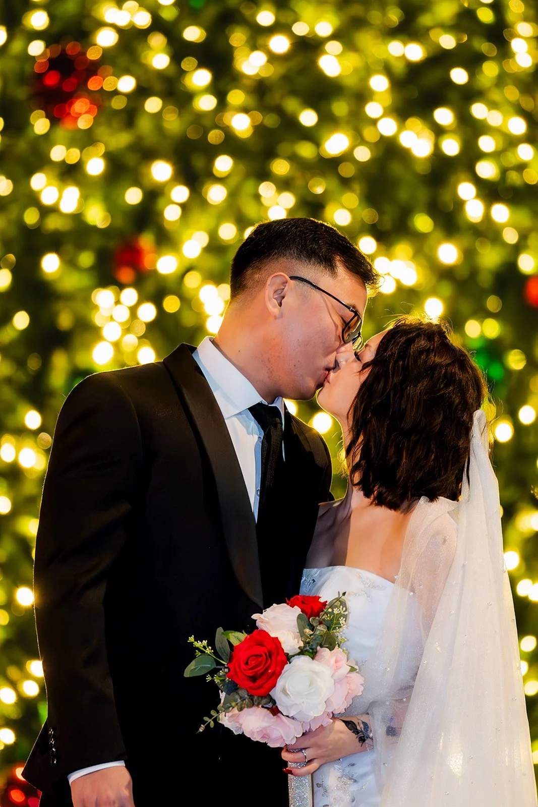 A newlywed couple shares a kiss in front of a decorated Christmas tree with golden lights, the groom wearing a black tuxedo and glasses, the bride in a white wedding dress holding a bouquet of red and white roses.