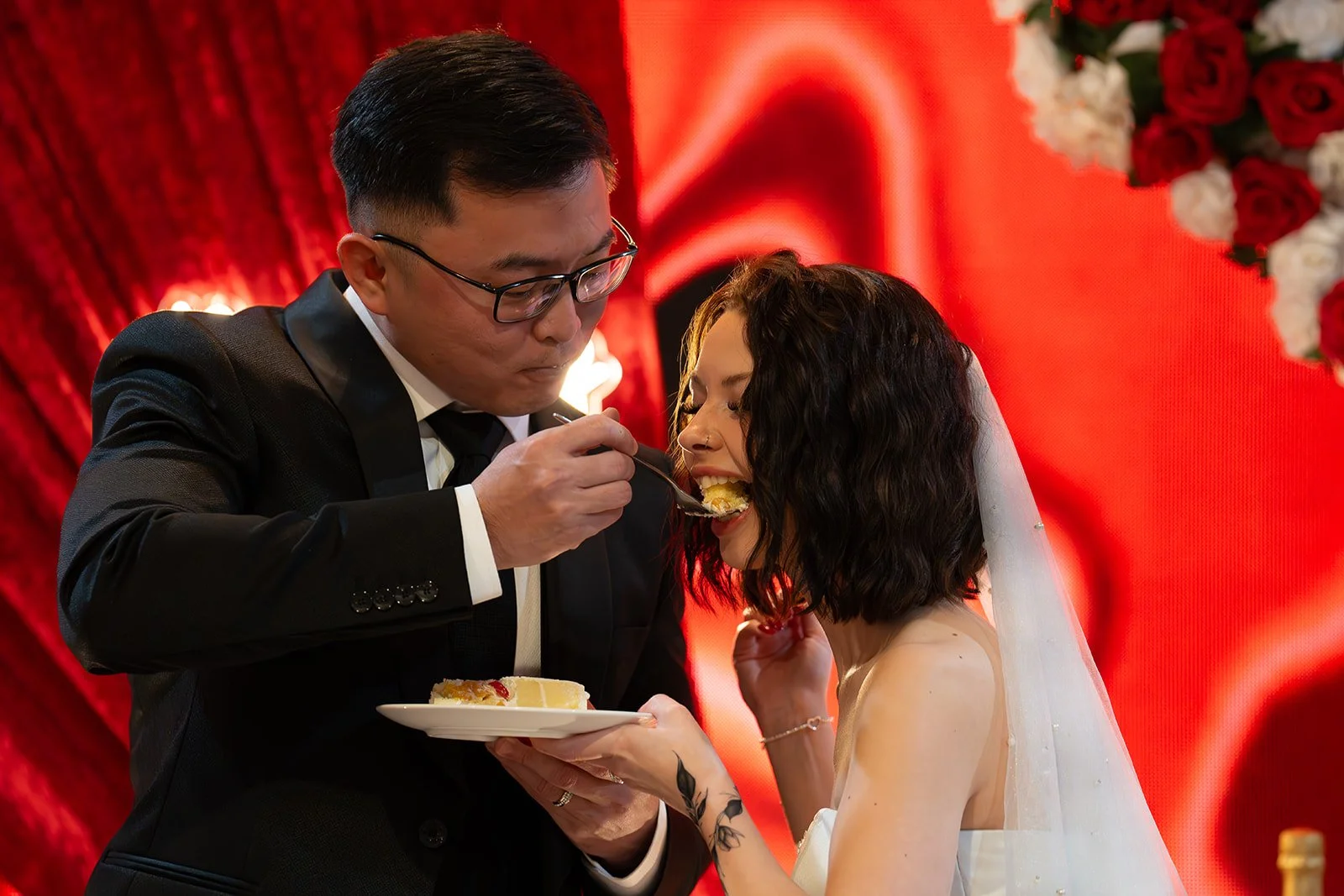 A groom in a black suit and glasses feeds cake to a bride with dark curly hair and tattoos on her arm, on her wedding day in front of a red decorated background with flowers.