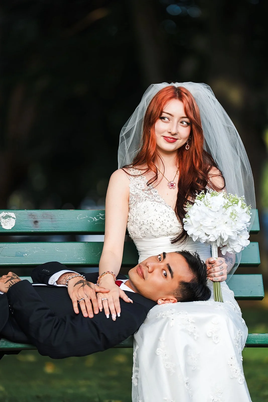 A bride with red hair, wearing a white wedding dress and veil, sitting on a green park bench. She is holding a bouquet of white flowers and looking slightly to her right. A groom with black hair, dressed in a black tuxedo, is lying down on the bench