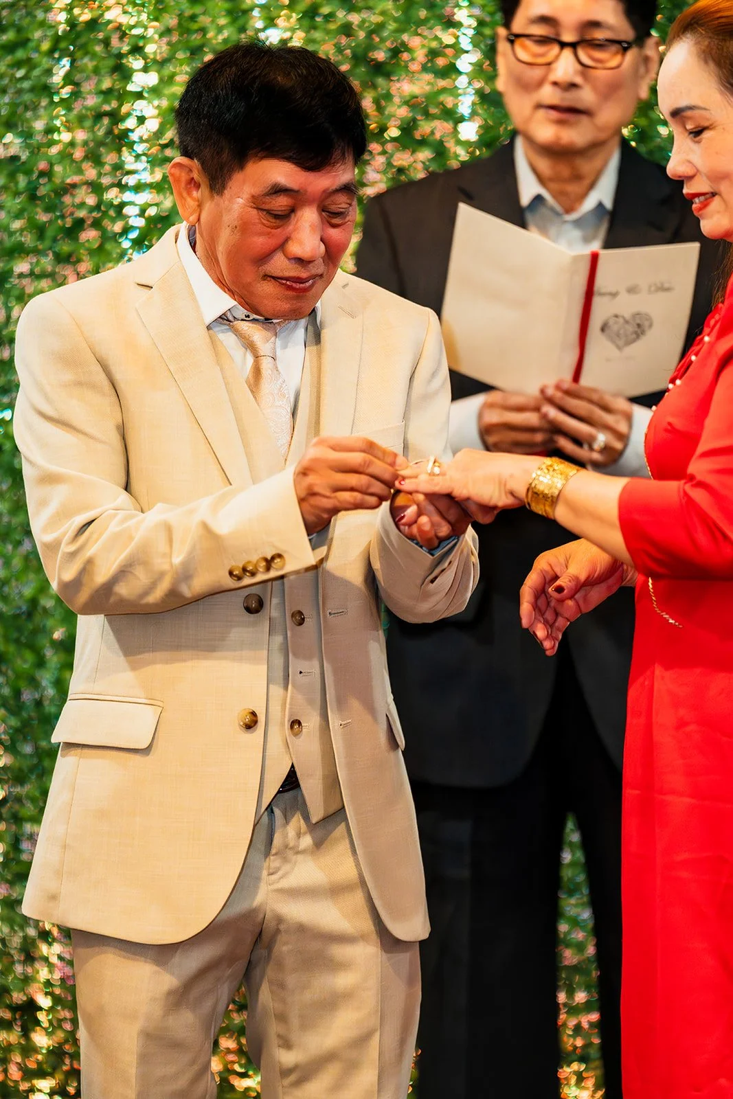 A man in a light-colored suit is placing a ring on a woman's finger during a wedding ceremony, with a man in a black suit reading from a book in the background, and a green, sparkly backdrop.