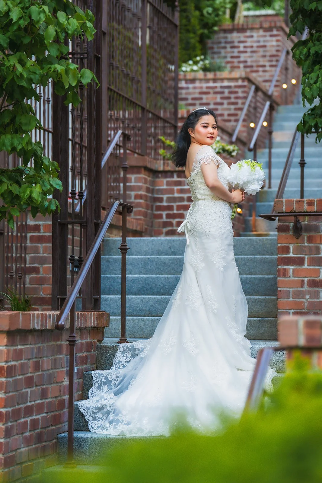 A woman in a wedding dress holding a bouquet of white flowers stands on outdoor steps near a brick building and greenery.
