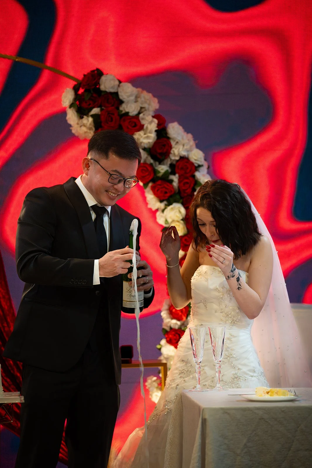 A bride and groom celebrating their wedding, with the groom pouring champagne and the bride looking at it, standing in front of a floral arch with red and white roses.