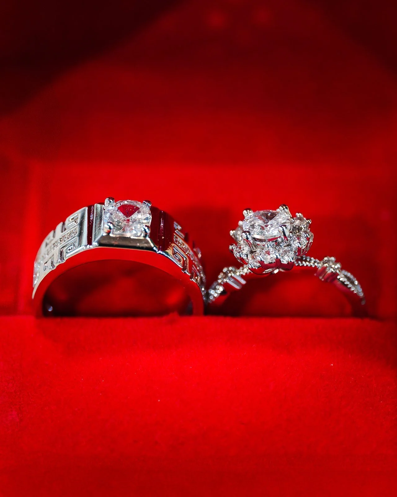 A pair of silver rings with diamonds, one with a square diamond and the other with a floral diamond design, set in a red velvet jewelry box.