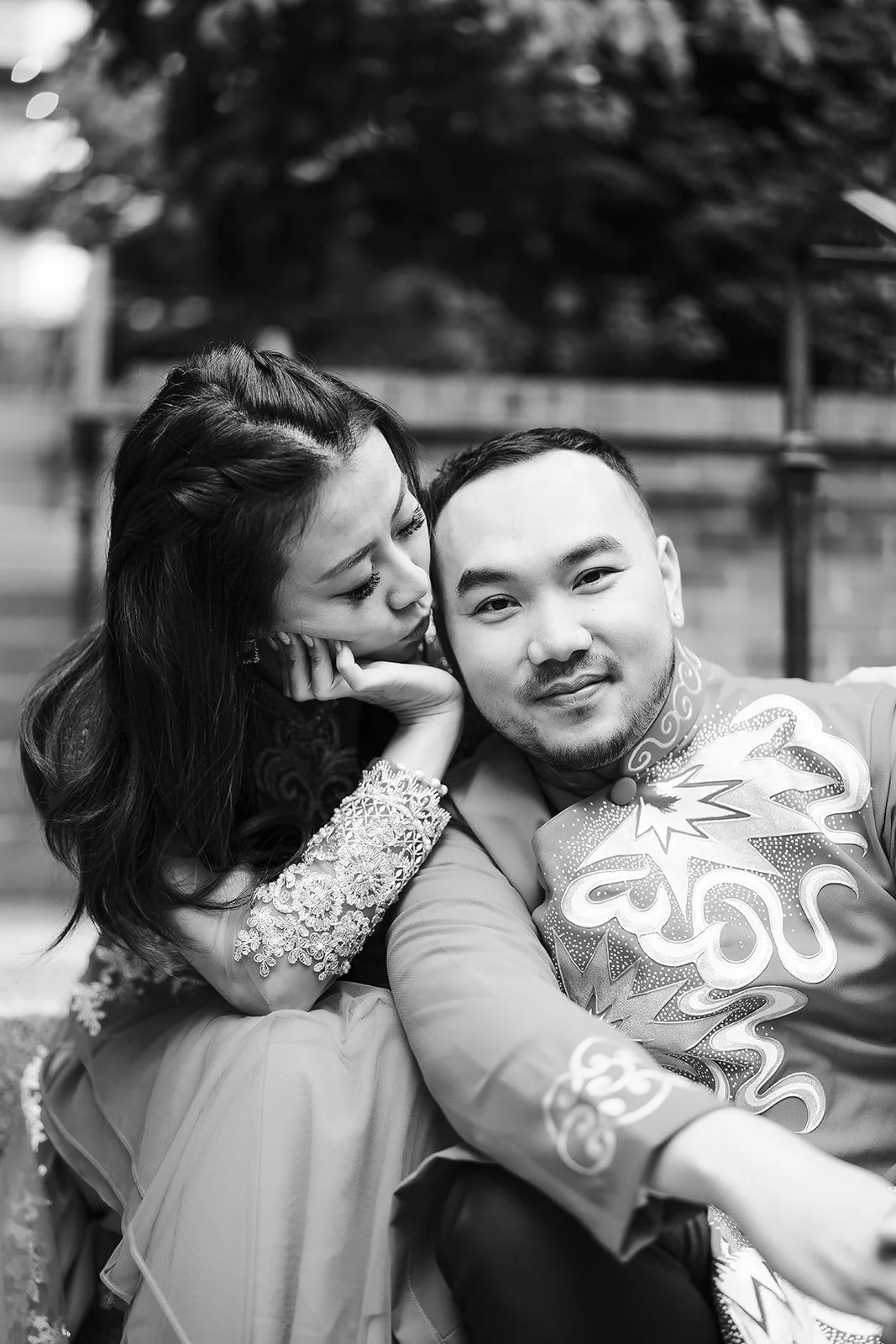 A couple taking a selfie outdoors, with the woman leaning her head on the man's shoulder, both smiling. The woman has long, dark hair and intricate lace sleeves. The man has short hair and is wearing a traditional patterned jacket.