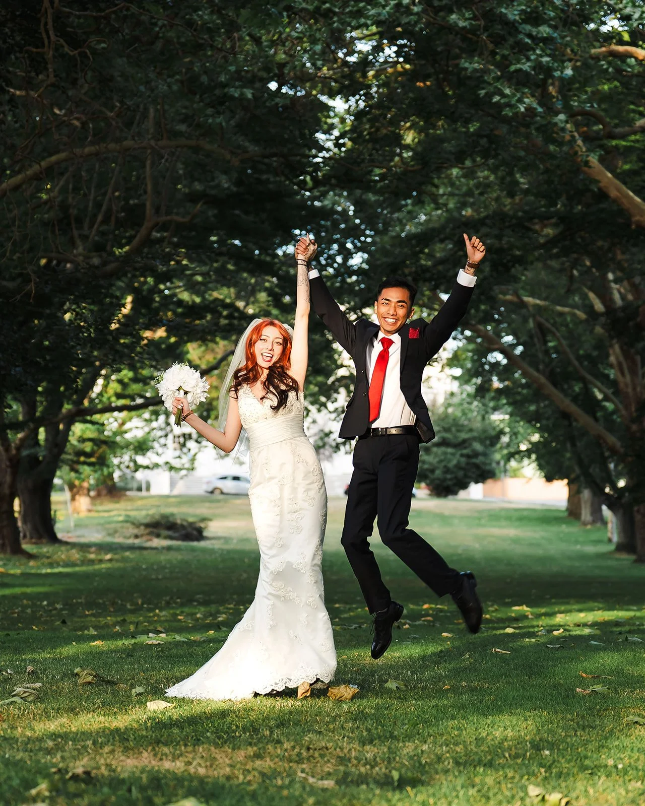 Happy bride and groom jumping in a park after their wedding, holding hands, with trees and green grass in the background.