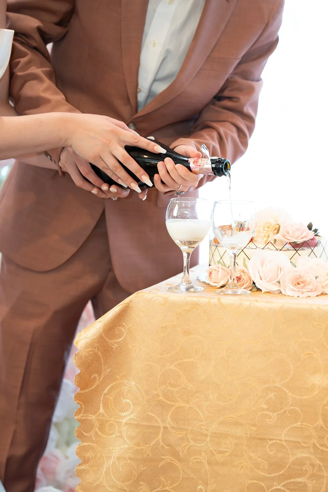 People pouring champagne into glasses during a celebration or wedding reception decorated with pink roses and a golden tablecloth.