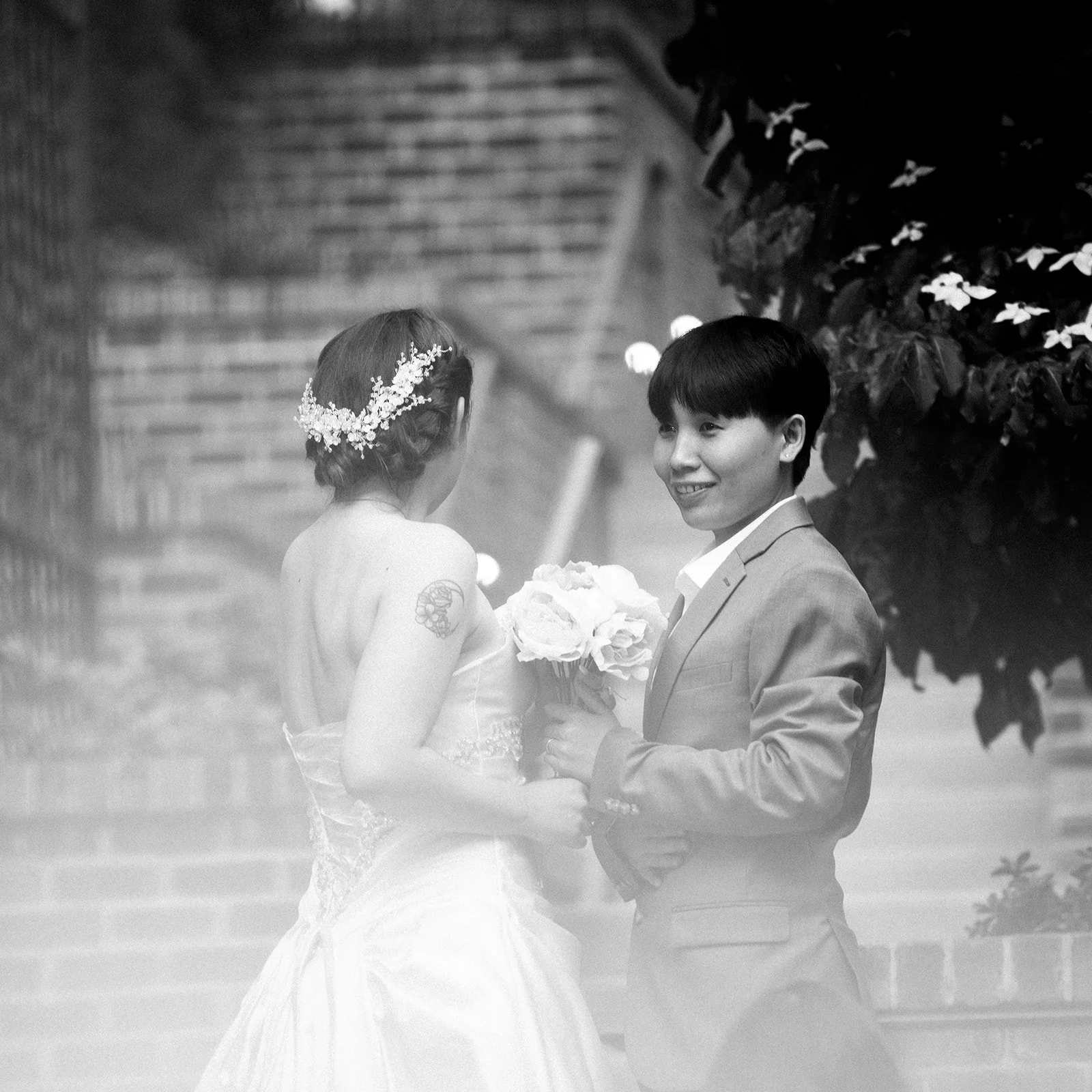 A black and white photo of a bride and groom standing outdoors, holding hands, with the groom smiling at the bride. The bride has a flower crown and a strapless wedding dress, while the groom is in a suit, holding a bouquet of flowers.