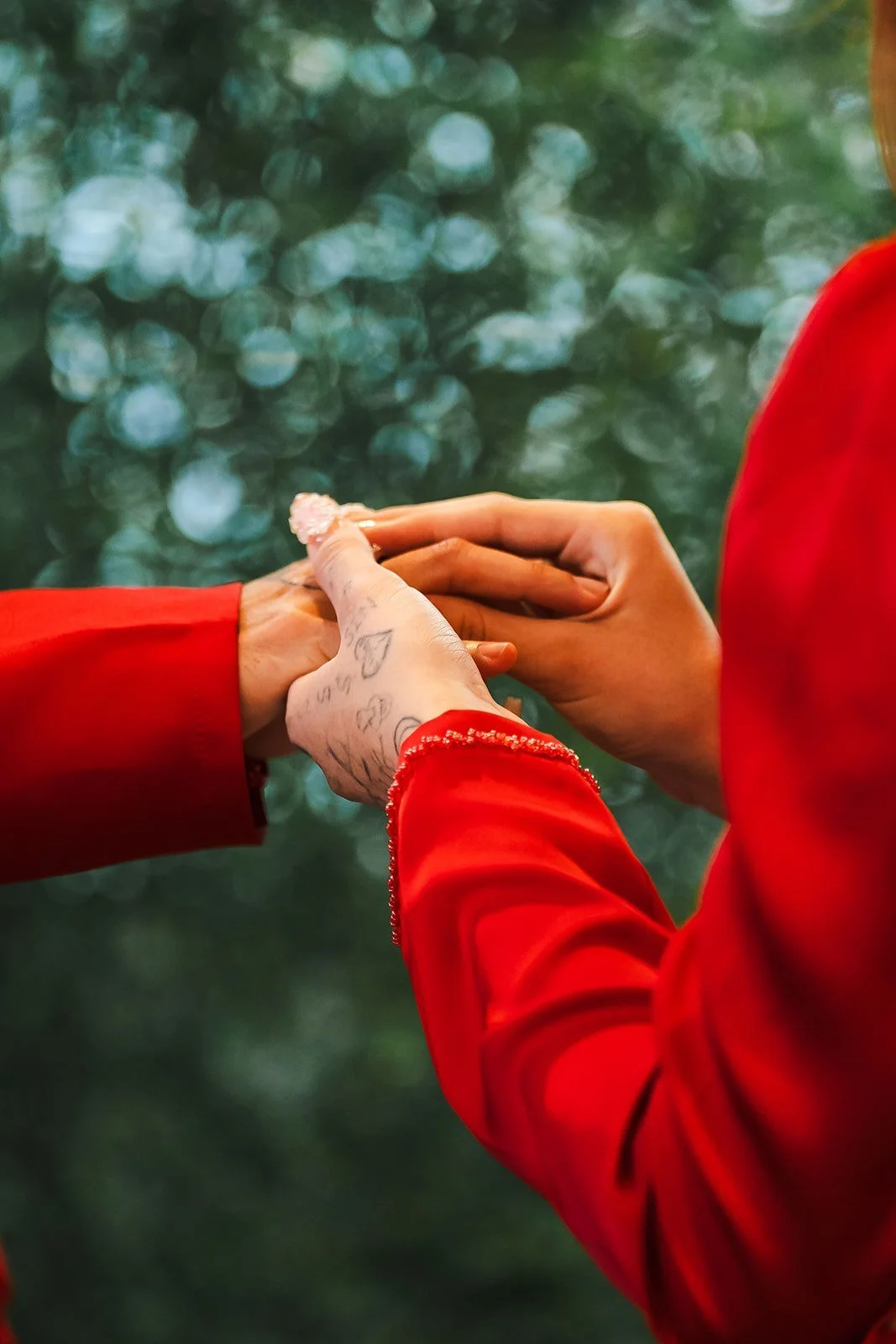 Two people holding hands, one person with tattoos and the other with smooth skin, both wearing red sleeves, against a blurred green background.