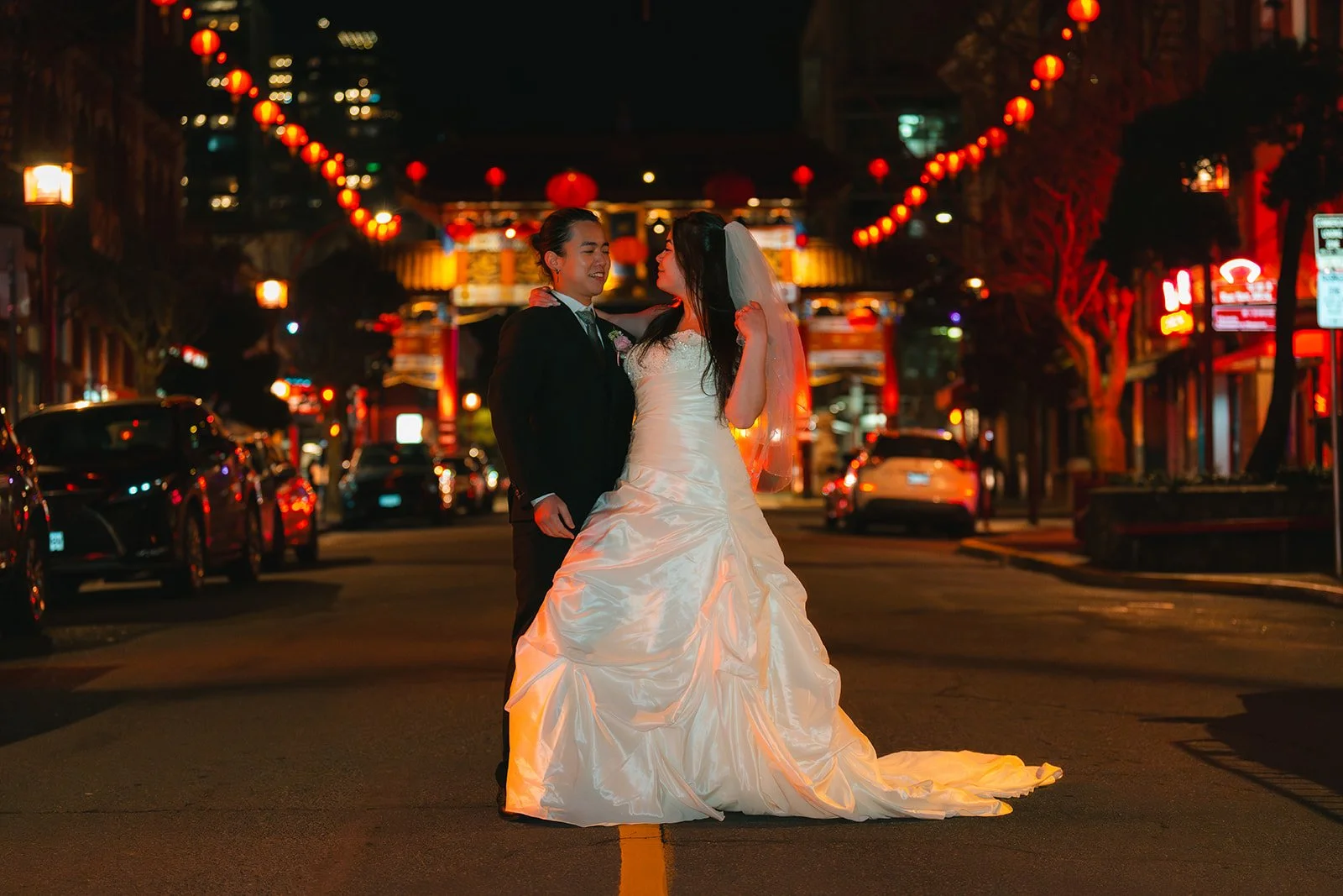 A bride and groom dancing in the middle of a street at night, decorated with red lanterns and illuminated signs, with cars parked along the sides.