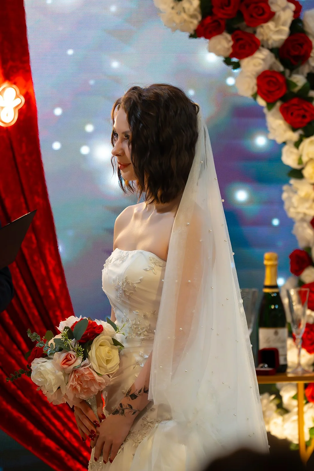 A bride in a white wedding dress holding a bouquet of roses and greenery, standing in front of a decorated backdrop with flowers and lights during a wedding ceremony.