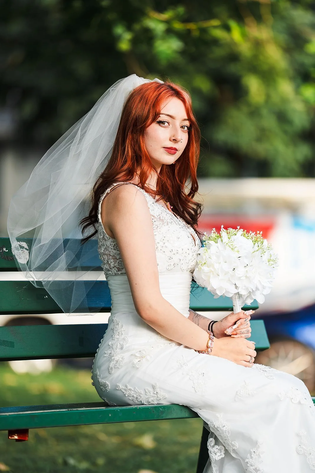 A bride with red hair and a white lace wedding dress sitting on a green park bench holding a bouquet of white flowers.