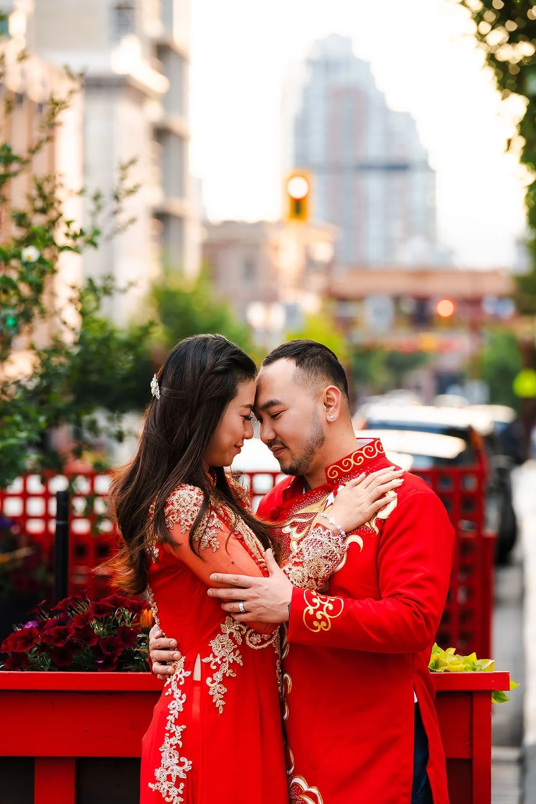 A couple in traditional Chinese wedding attire embrace outdoors, gazing into each other's eyes, with tall buildings and greenery in the background.