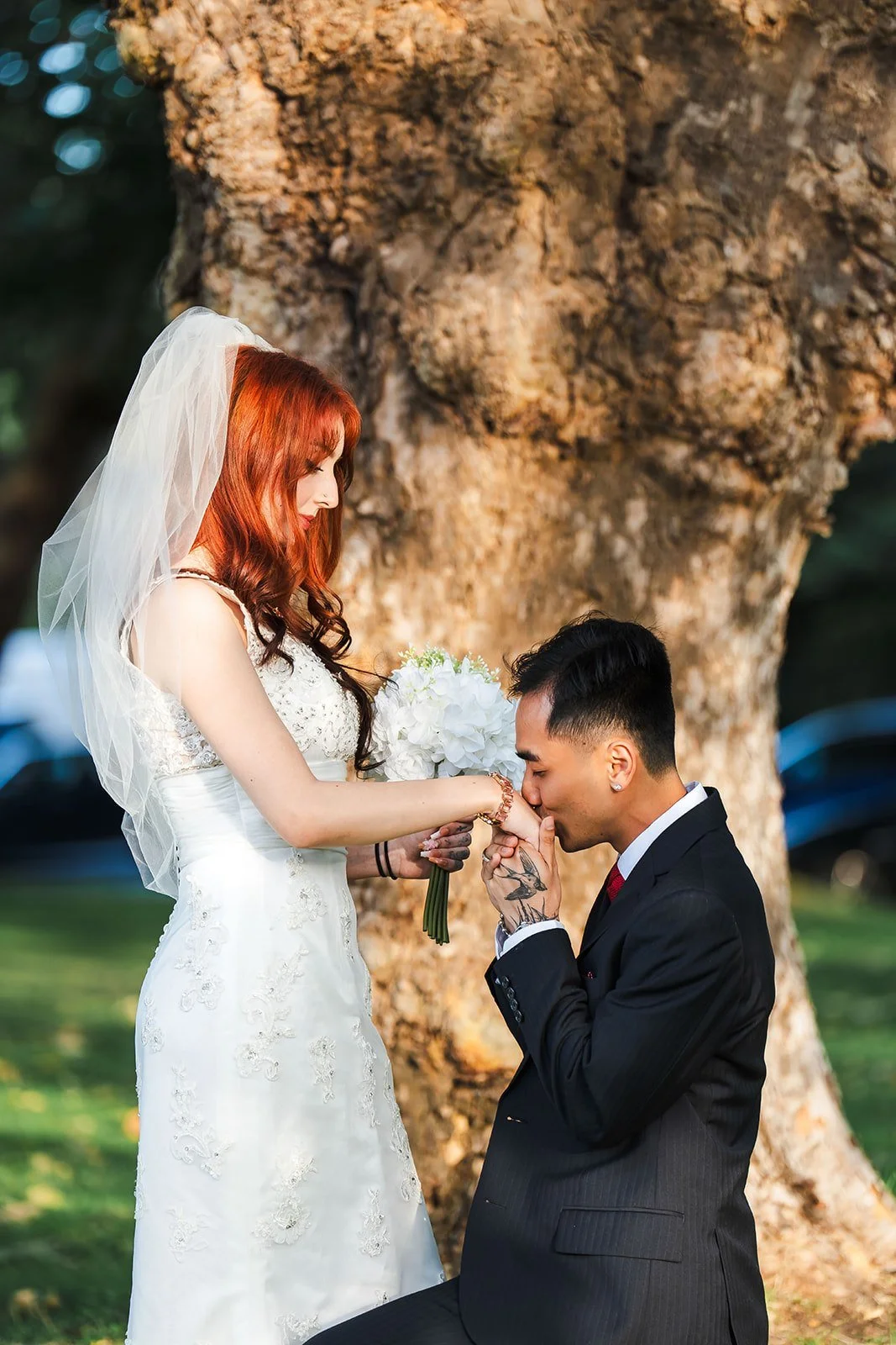 A bride with red hair wearing a white wedding dress and veil is holding a bouquet of white flowers. A groom with black hair in a black suit is kneeling and kissing her hand in front of a large tree outdoors during daytime.
