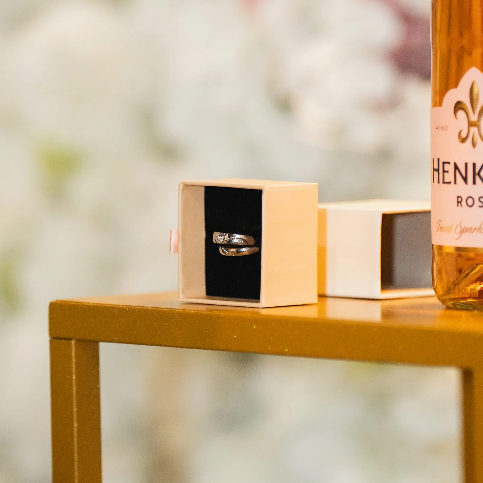 A silver ring with a small gemstone in a white box with a black interior, and a bottle of rosé wine on a gold-colored table.