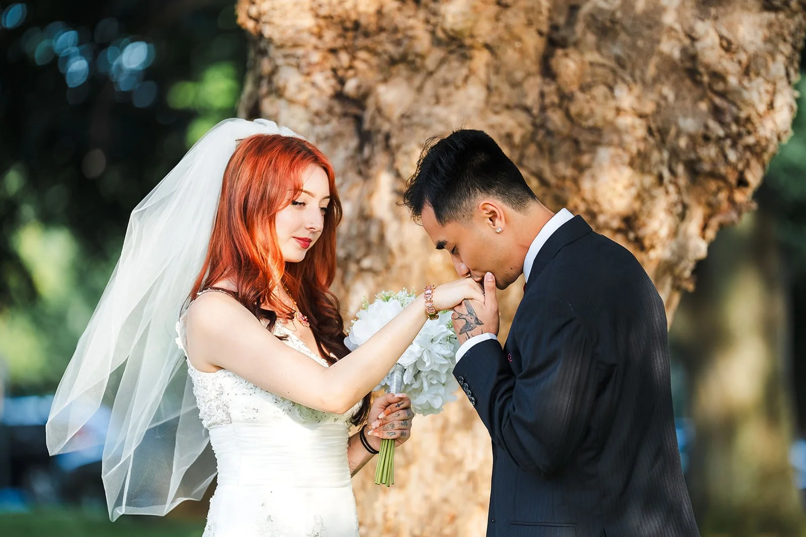 A bride with red hair and a veil holding a bouquet of white flowers, and a groom in a black suit with short black hair, in a park setting during a wedding ceremony.