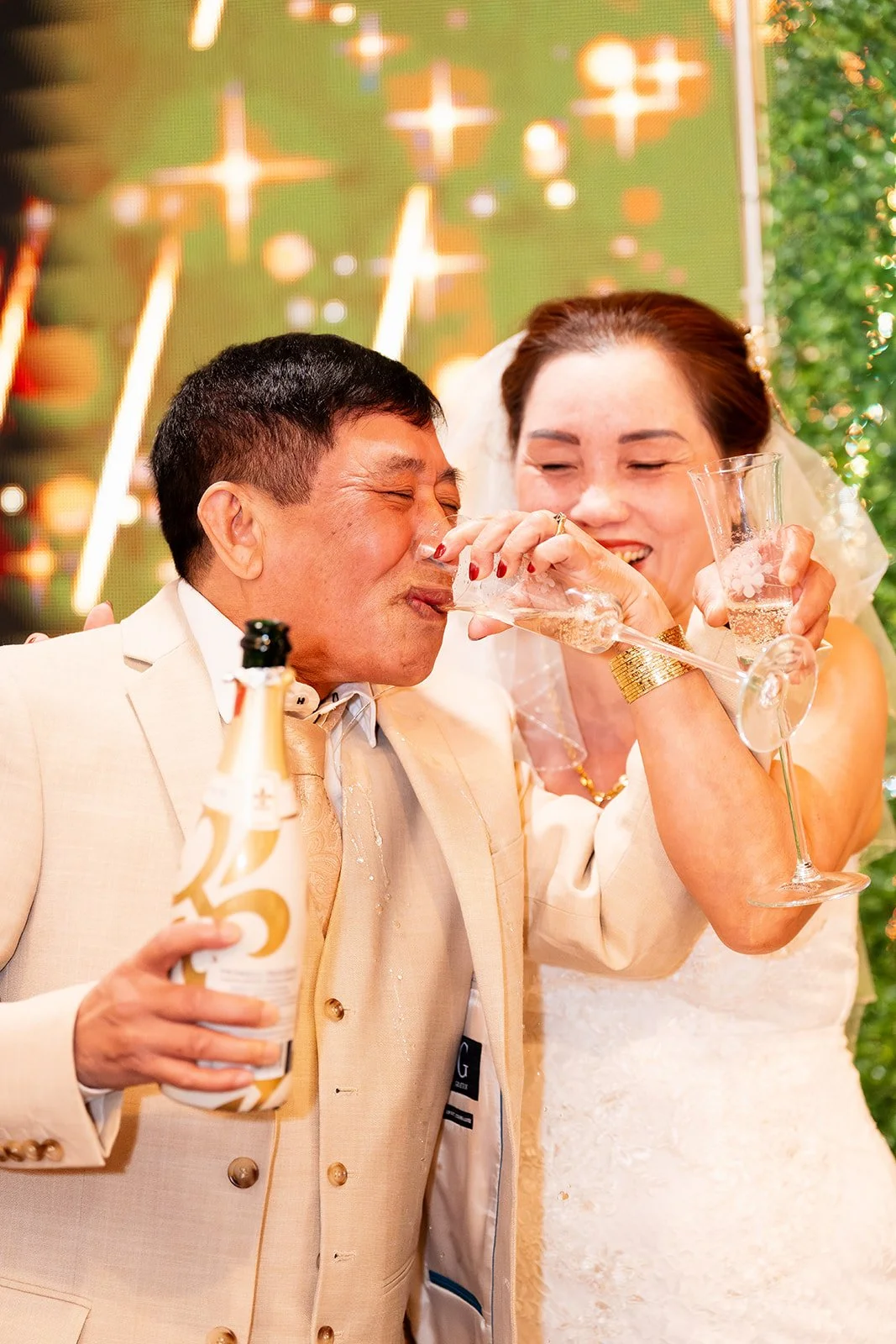 A woman in a wedding dress and a man in a beige suit celebrating with drinks, smiling and laughing, at a wedding reception.