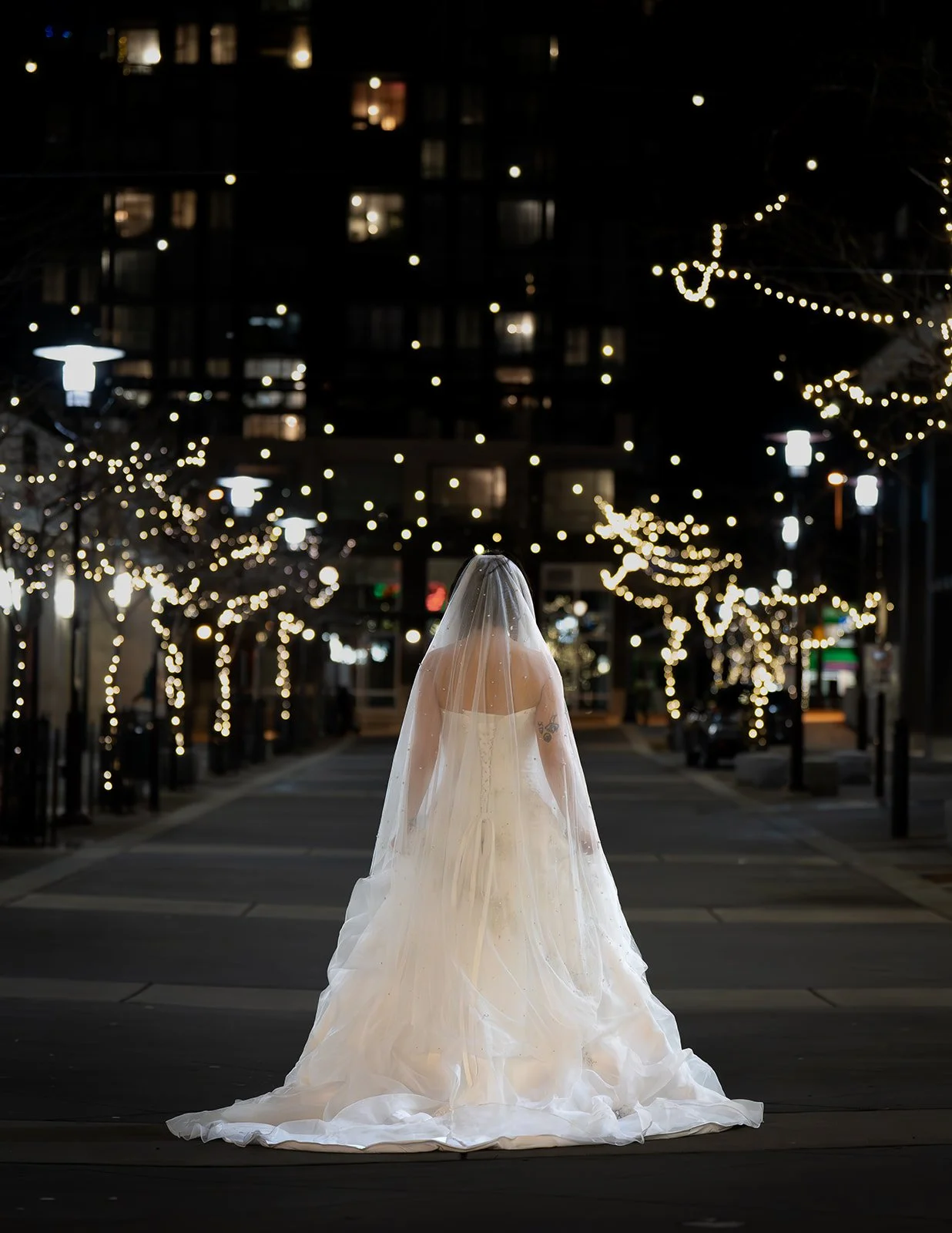 A person in a wedding dress with a veil standing alone in a lit city street at night, decorated with string lights and illuminated trees.