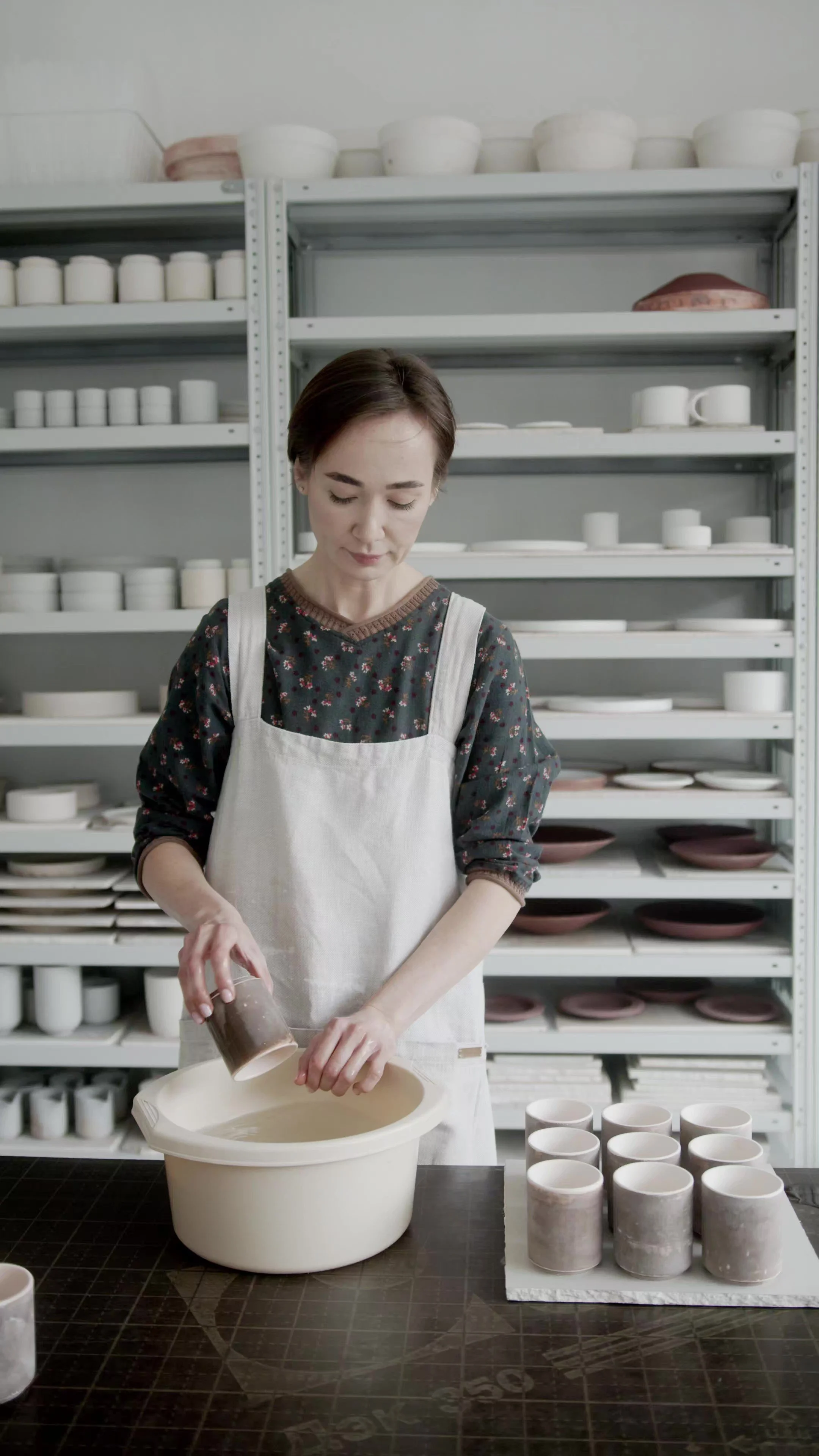 A woman in an apron is working on a pottery project in a studio, pouring a brown slip into a ceramic piece.