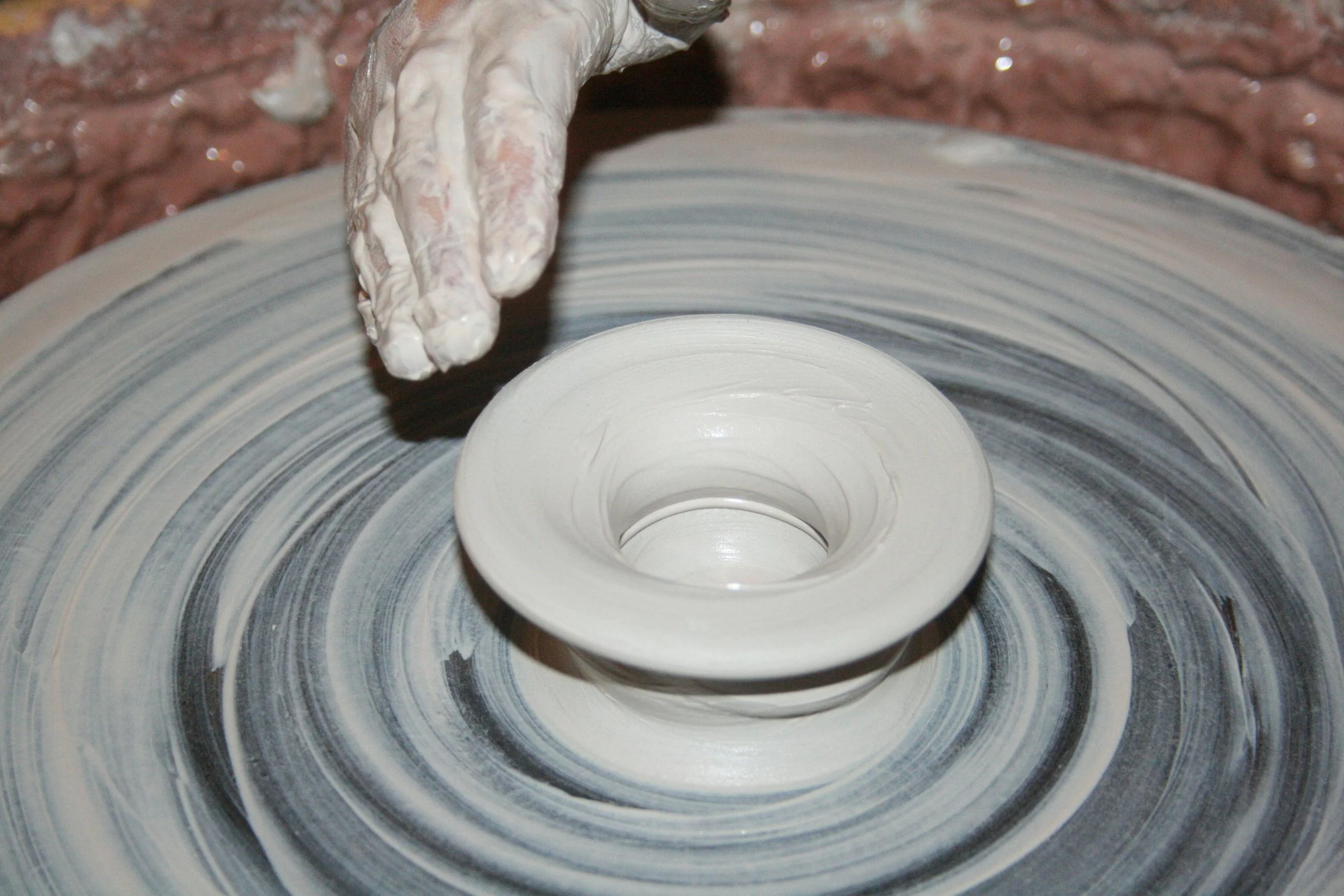 A person shaping pottery on a wheel, wearing a white apron, with a brick wall in the background.
