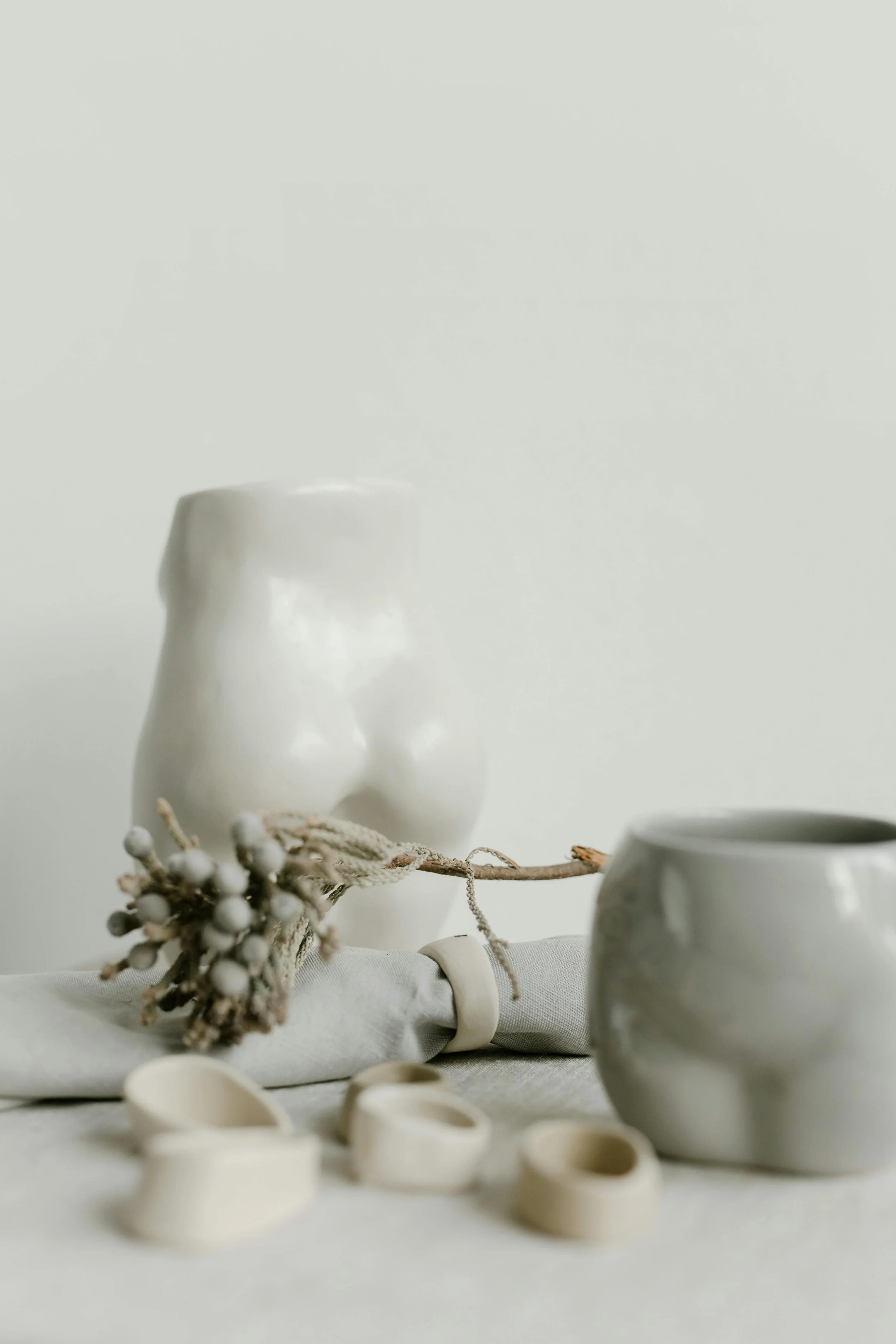 Close-up of a white ceramic sculpture of a small cocoon, with a branch of dried berries and a decorative ribbon in the foreground. The background features additional white ceramic objects, creating a minimalist aesthetic.