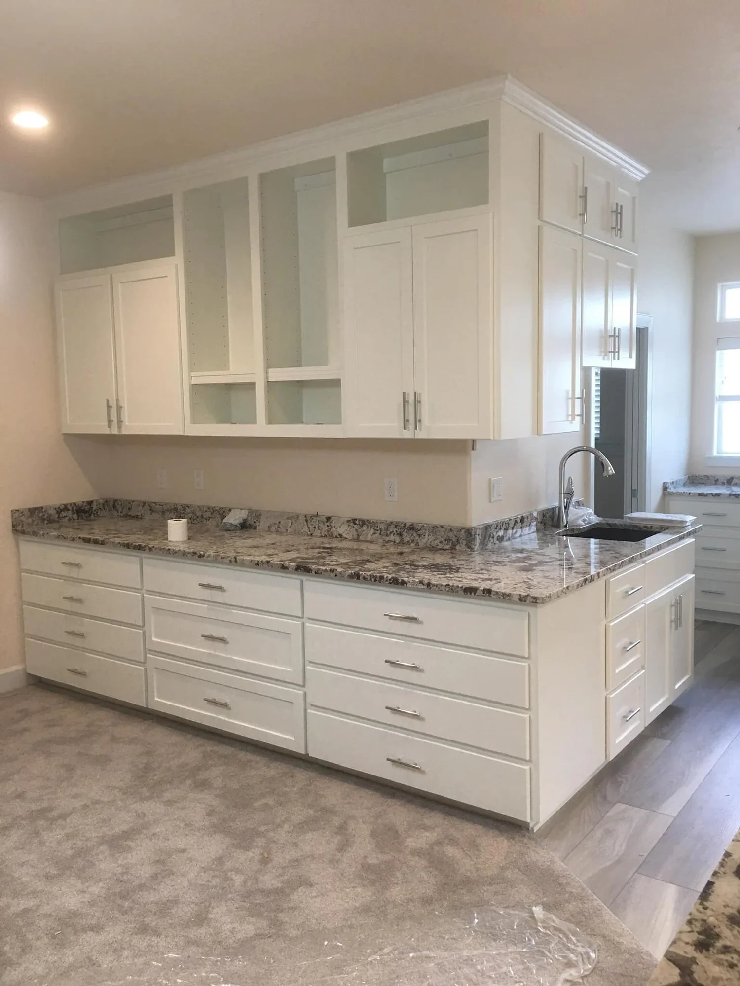 A kitchen with white cabinets, granite countertops, a sink, and a window in the background. The upper cabinets are open with no doors on some sections.