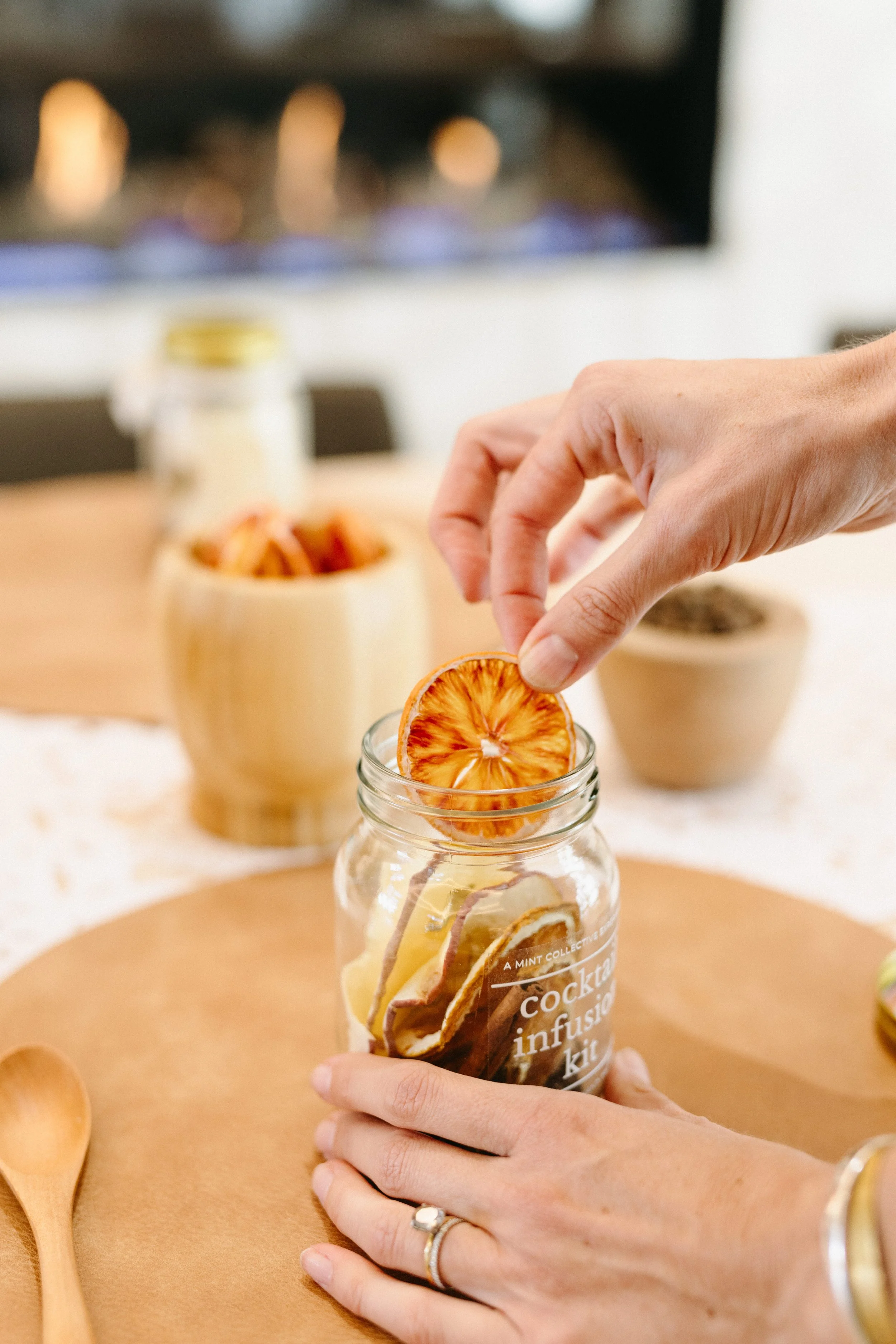 Hands assembling a DIY craft cocktail infusion kit with dried fruits and botanicals in a glass jar.