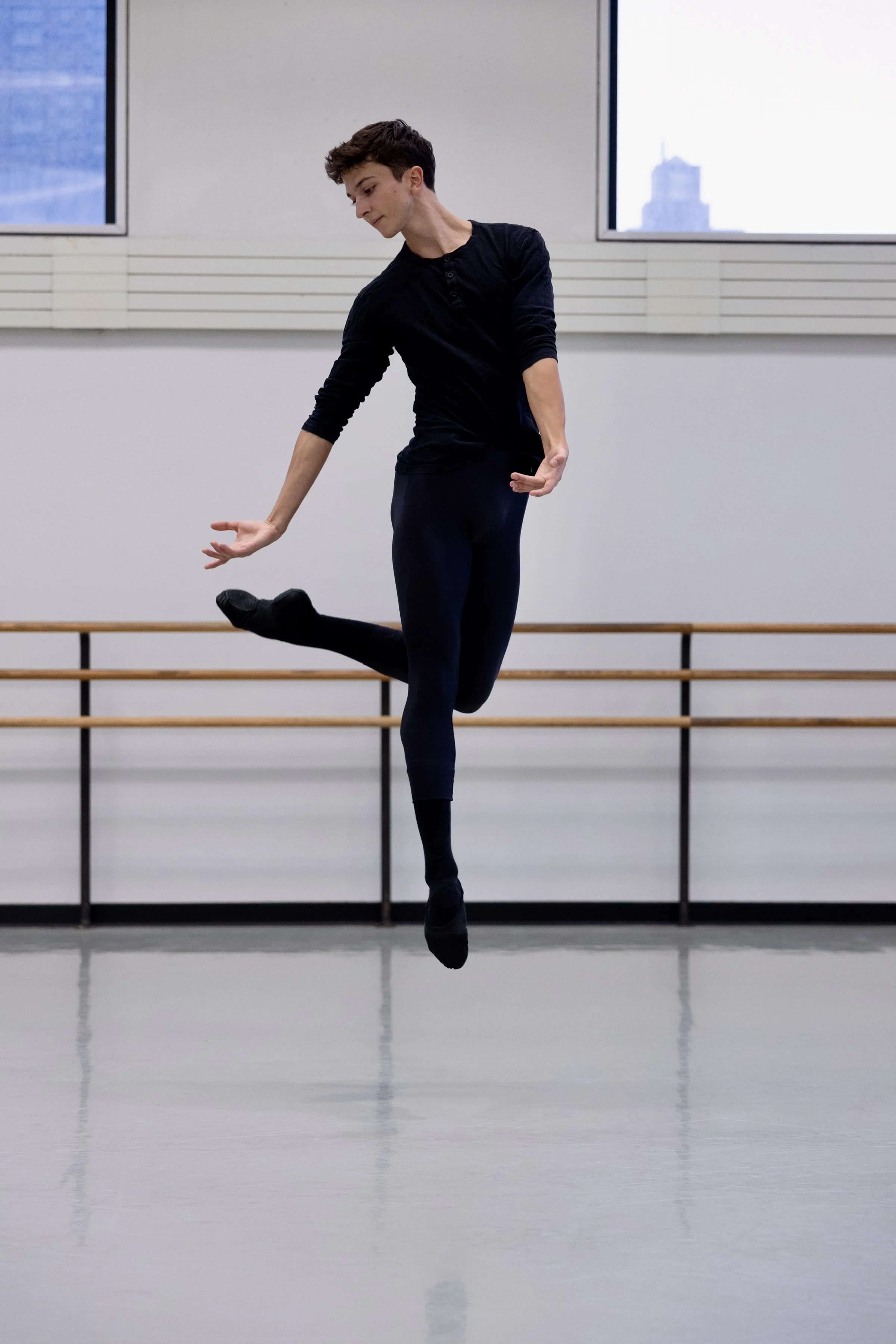 A young male dancer practicing ballet in a dance studio, mid-jump with one leg lifted and arms extended, wearing black workout clothes New York City Lincoln Center.