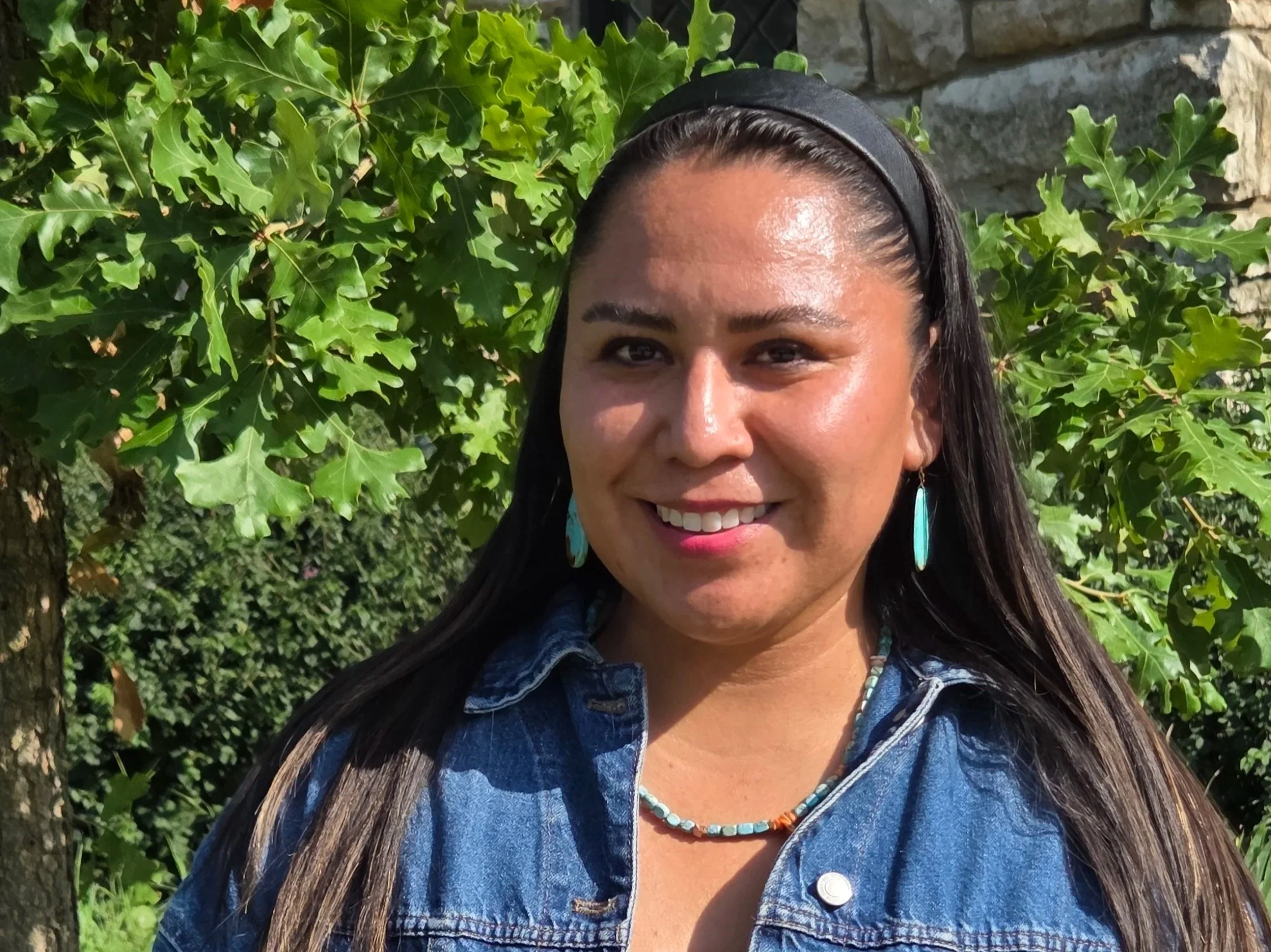 Outdoor portrait of a smiling woman with long dark hair, wearing a denim jacket, turquoise jewelry, and a mosaic-patterned dress.