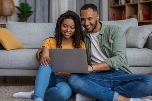 A smiling couple sitting on the floor in front of a gray sofa, looking at a laptop together in a cozy living room.