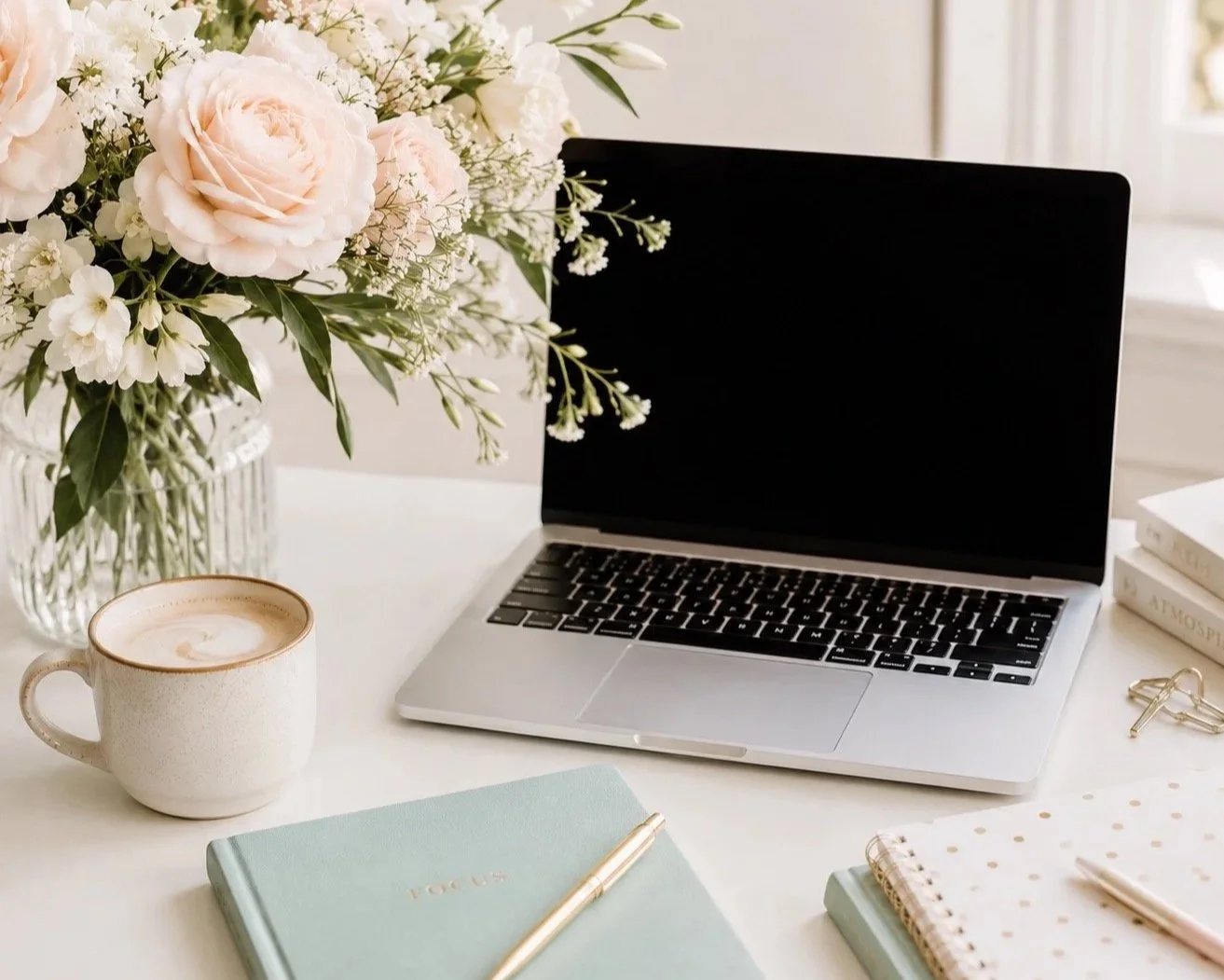 A workspace with a bouquet of flowers, an open laptop, a coffee mug, notebooks, a gold pen, and gold paper clips on a white desk.
