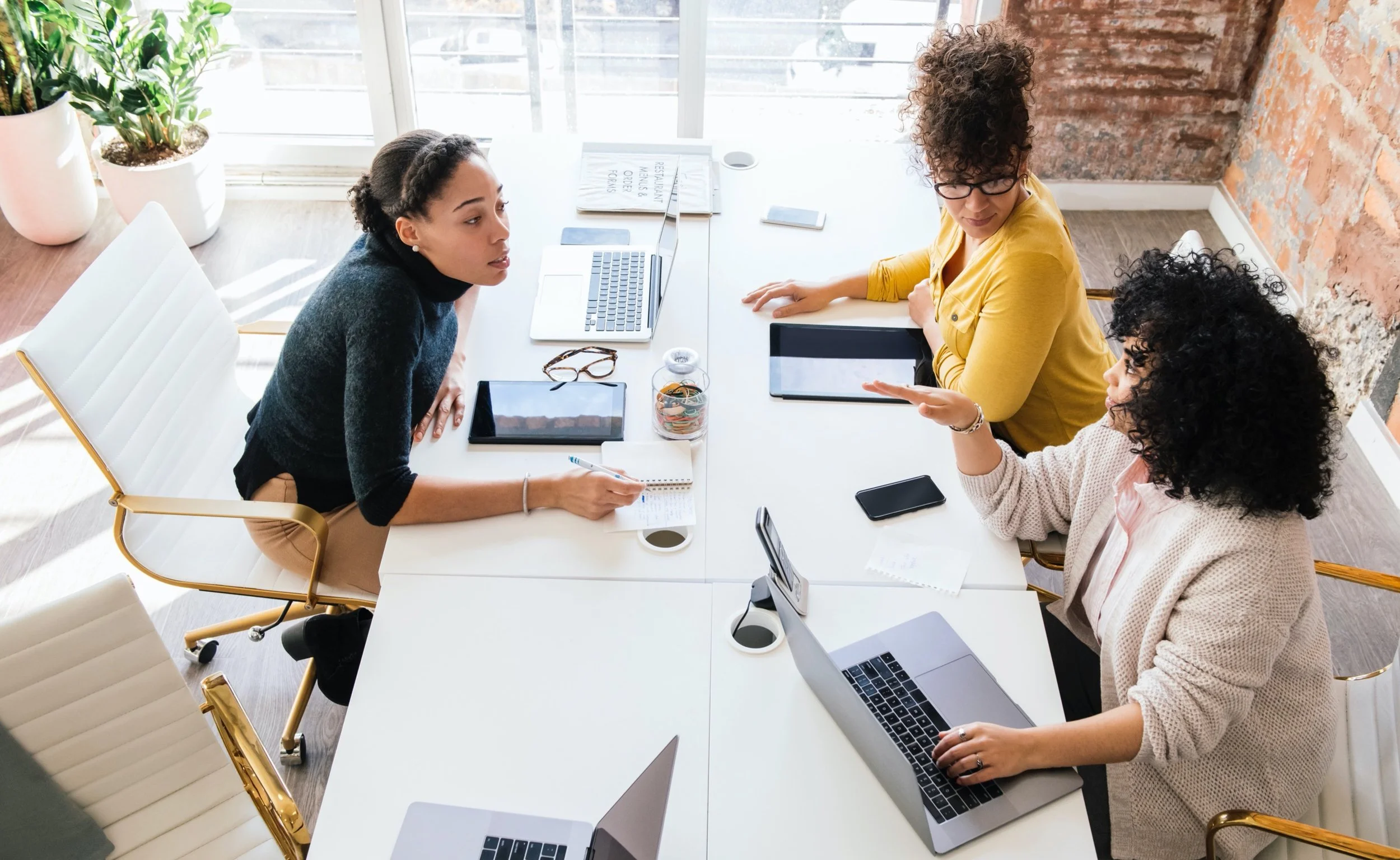 Four women in a meeting room with laptops, tablets, and notebooks, engaged in discussion around a white conference table near large windows.