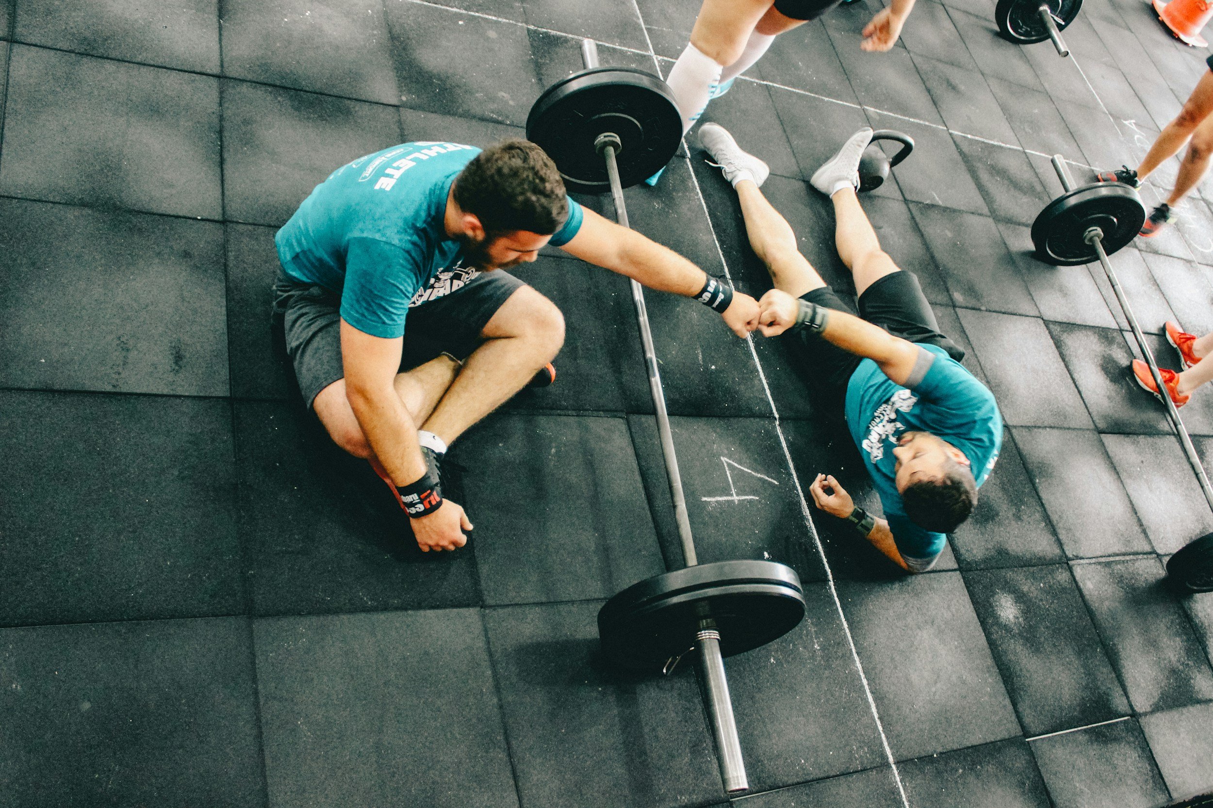 A high-angle photograph looking down at two fit men fist-bumping while lying on the floor of a gym after a workout, representing client bookings for a fitness company.
