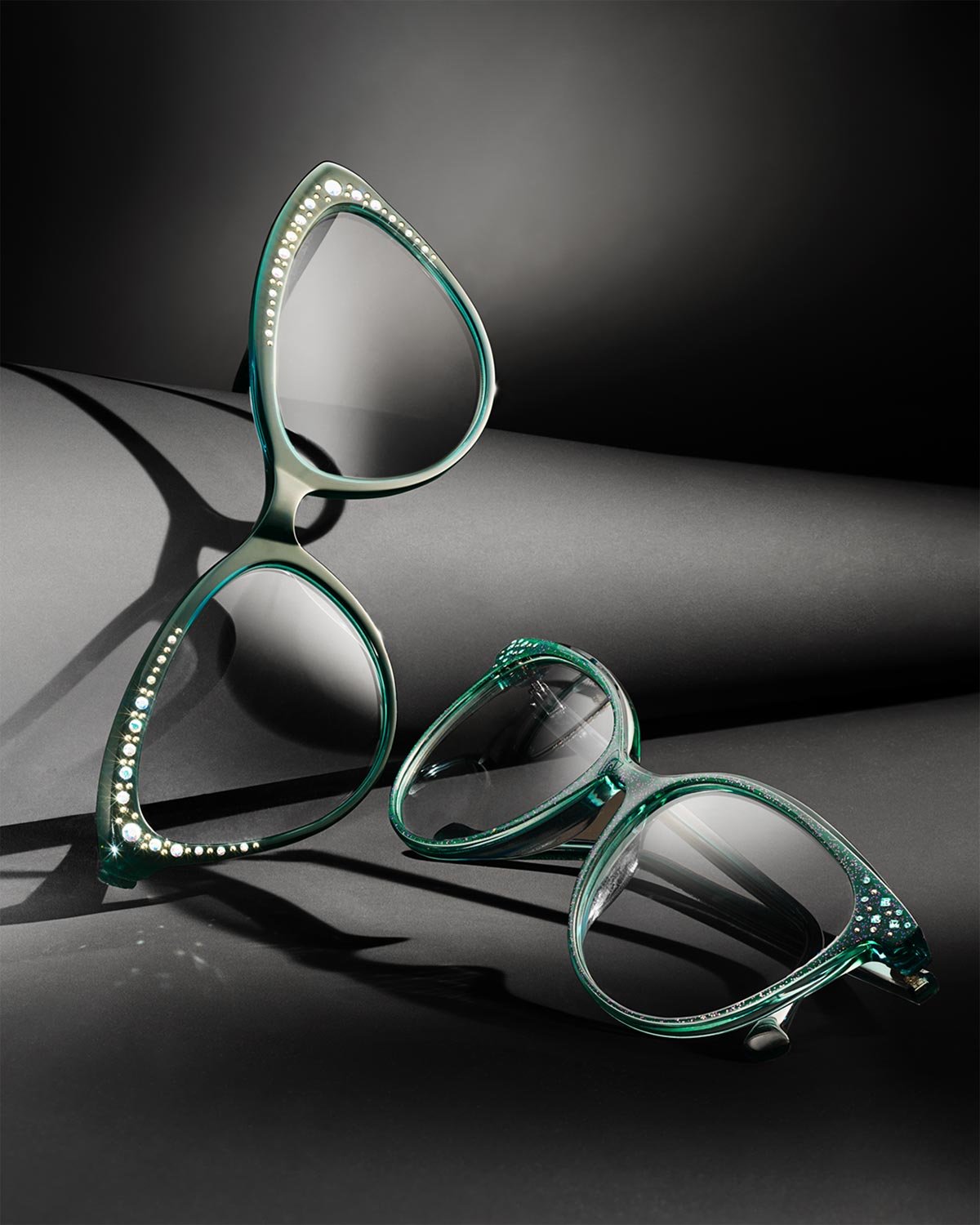 Two pairs of stylish sunglasses with embellished frames, one standing upright and the other lying down, on a dark surface with dramatic lighting and shadows.