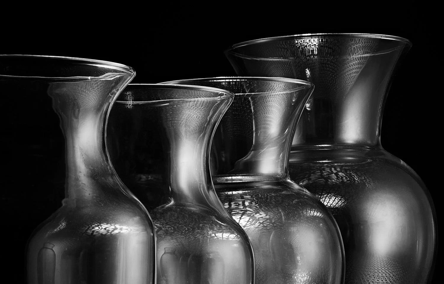 Close-up of four transparent glass vases with curved necks, set against a black background, overlapping each other.