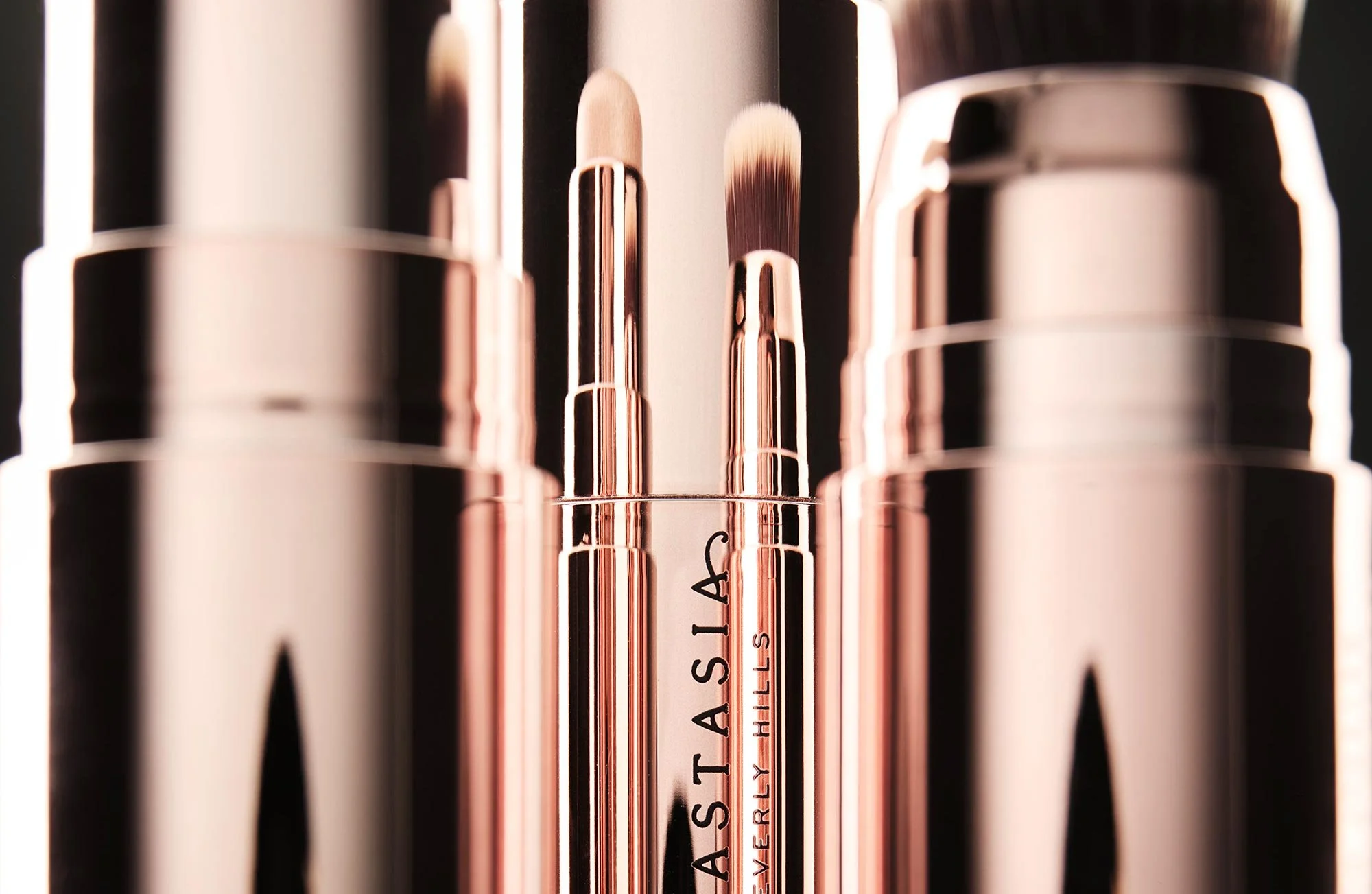 Close-up of various makeup brushes with rose-gold handles, standing upright on a reflective surface.