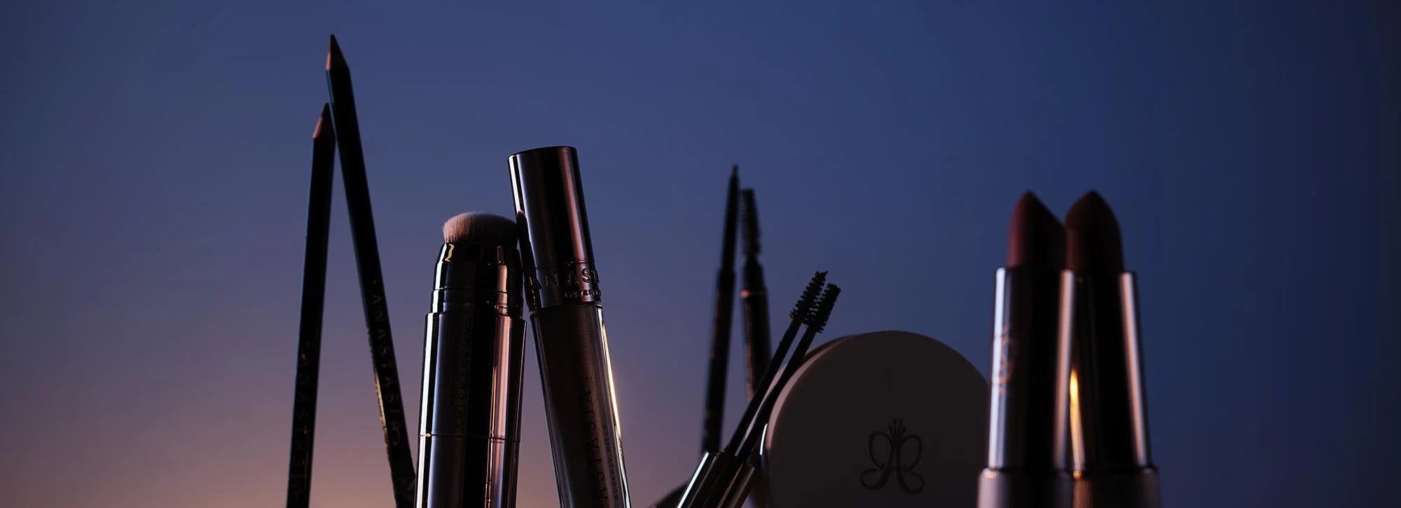 Various makeup products including lipsticks, mascaras, eyebrow pencils, and brushes on a dark background with subtle lighting.