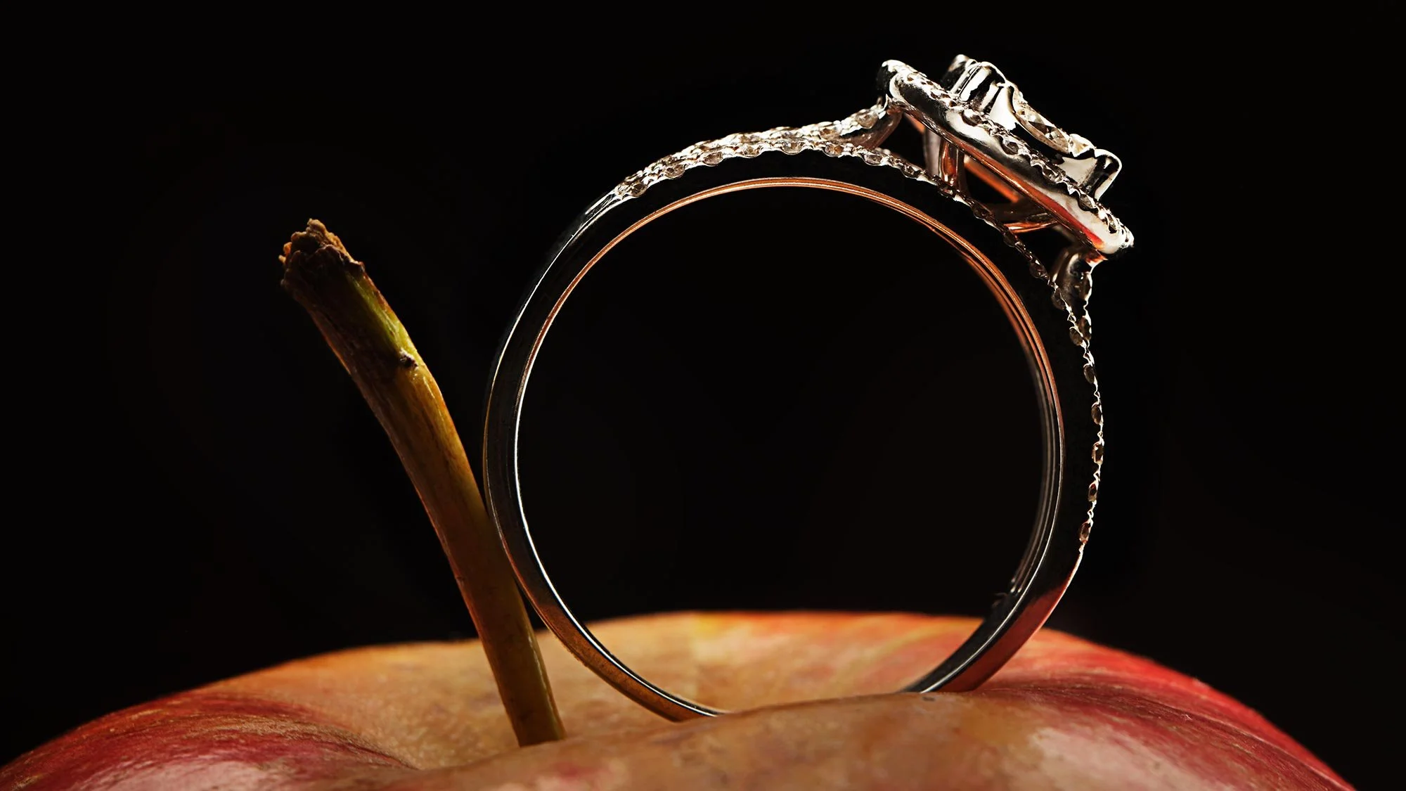 A silver ring with a stone on top, placed on a red apple with a brown stem, against a black background.