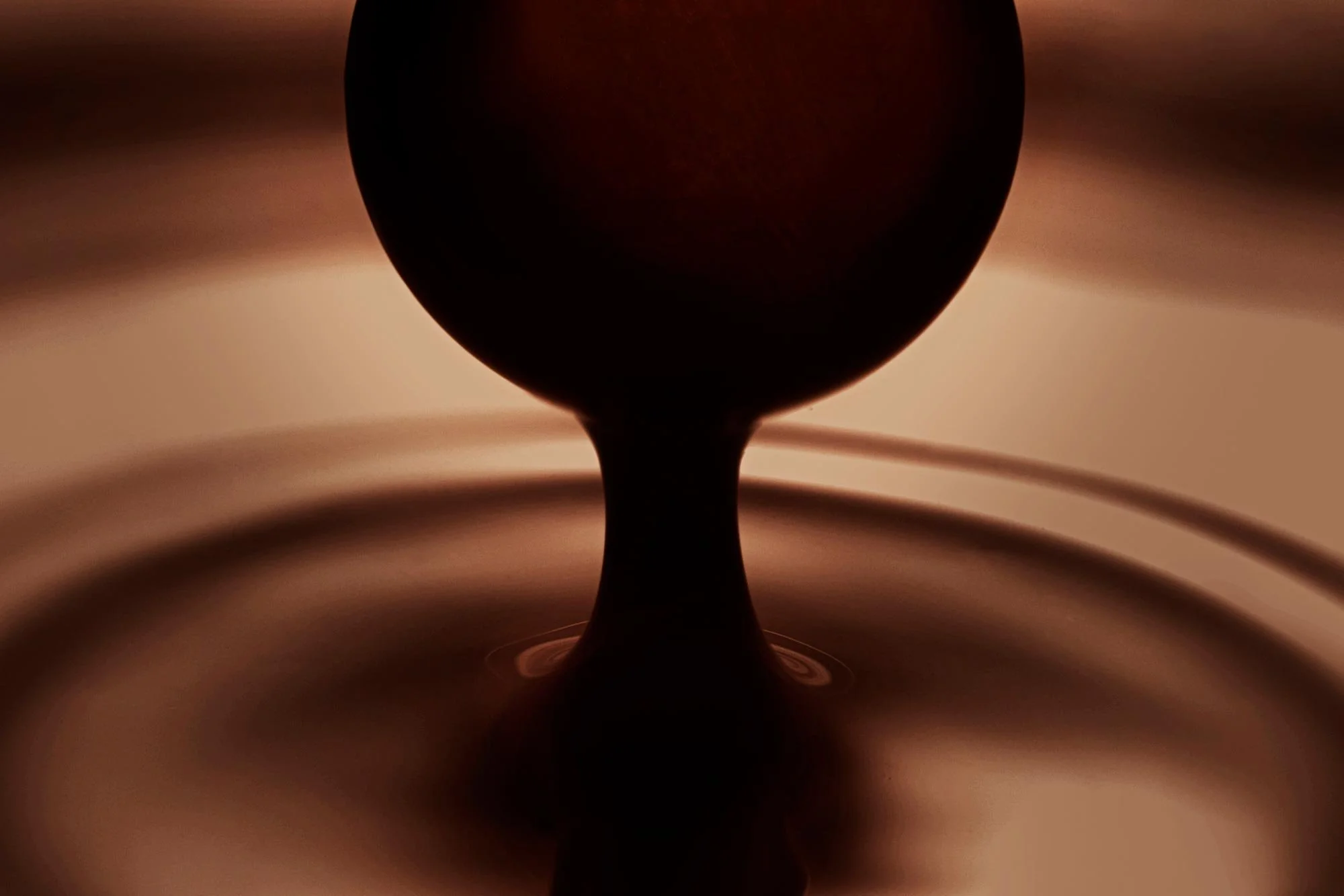 Close-up of a dark chocolate droplet suspended above a liquid surface, creating ripples in a warm, gradient background.