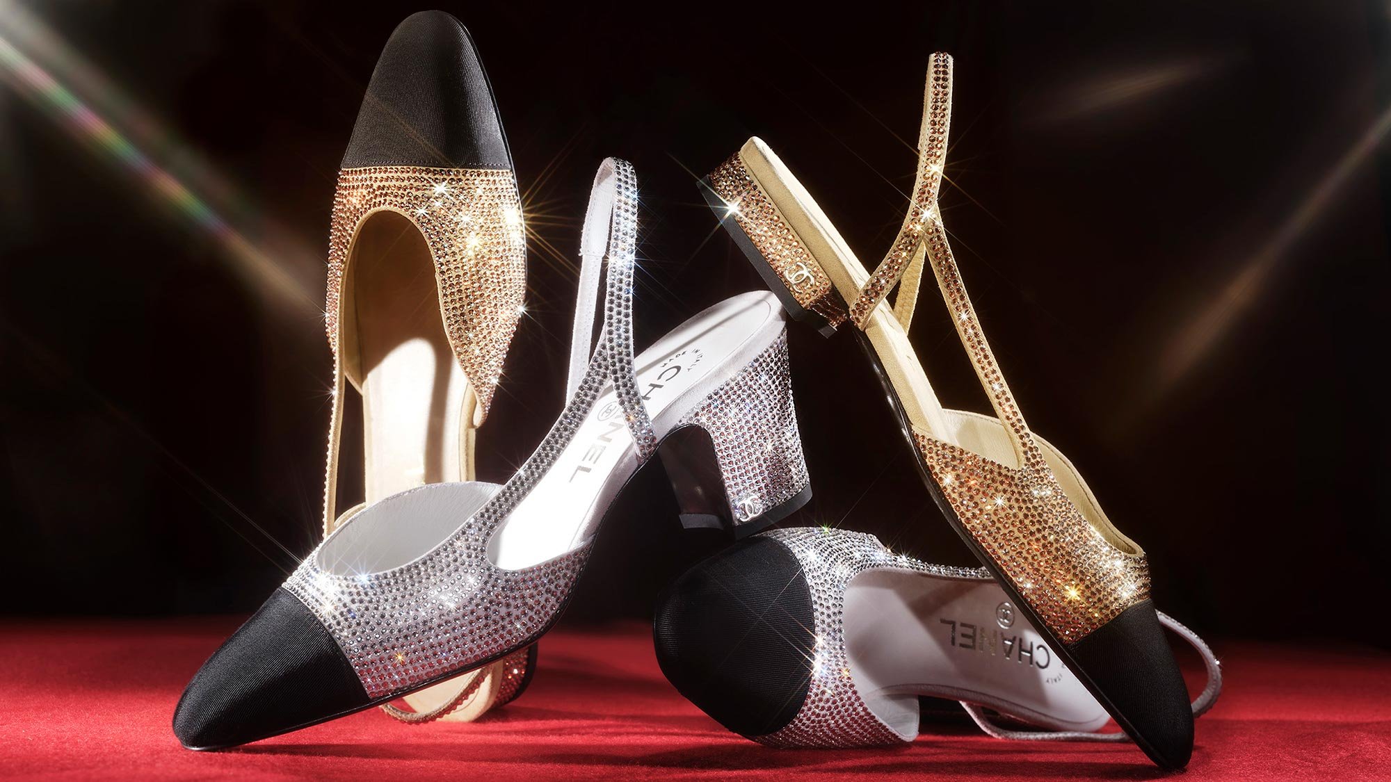 Two pairs of high-heeled shoes decorated with rhinestones, one black and one gold, arranged on a red carpet with a dark background.