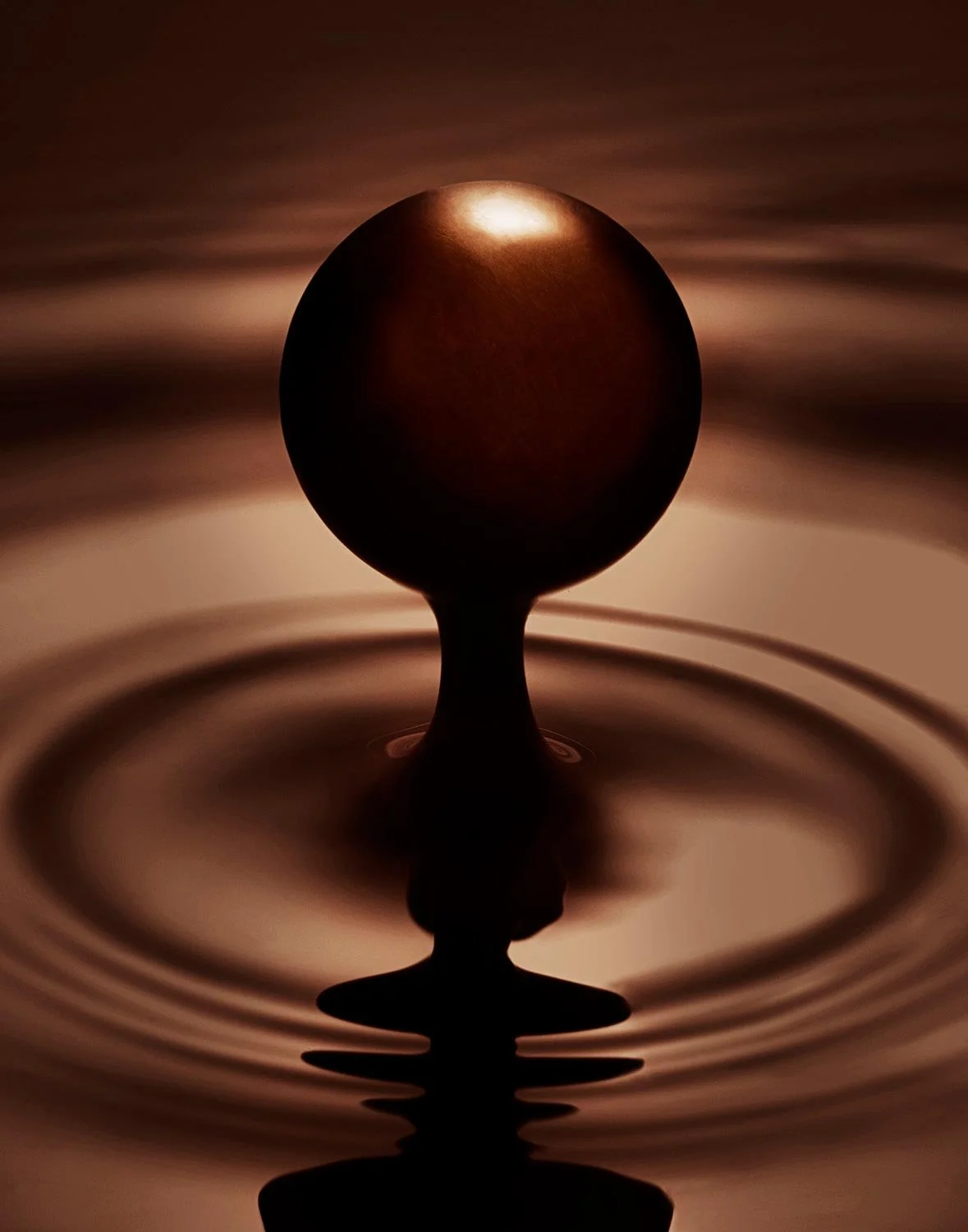 Close-up of a chocolate droplet creating ripples on a chocolate liquid, with a dark chocolate ball suspended above.