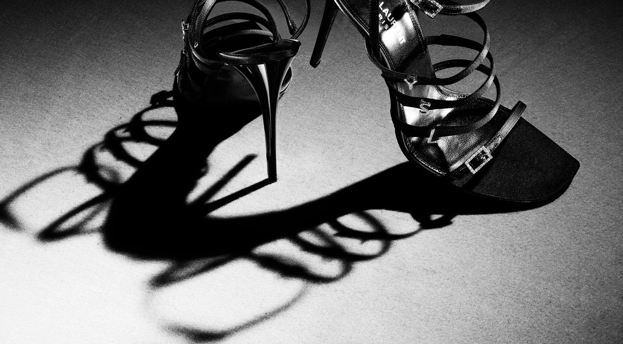 Black high-heeled sandal with multiple straps and a buckle, casting a complex shadow on a grey surface.