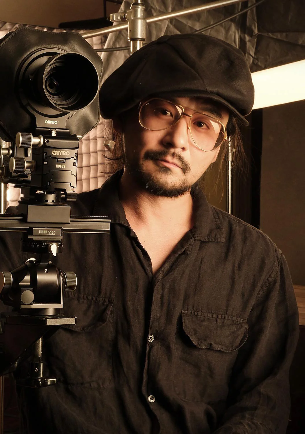 A man with long hair, glasses, and a beard wearing a black beret and black shirt, standing next to a professional camera on a tripod in a studio setting.