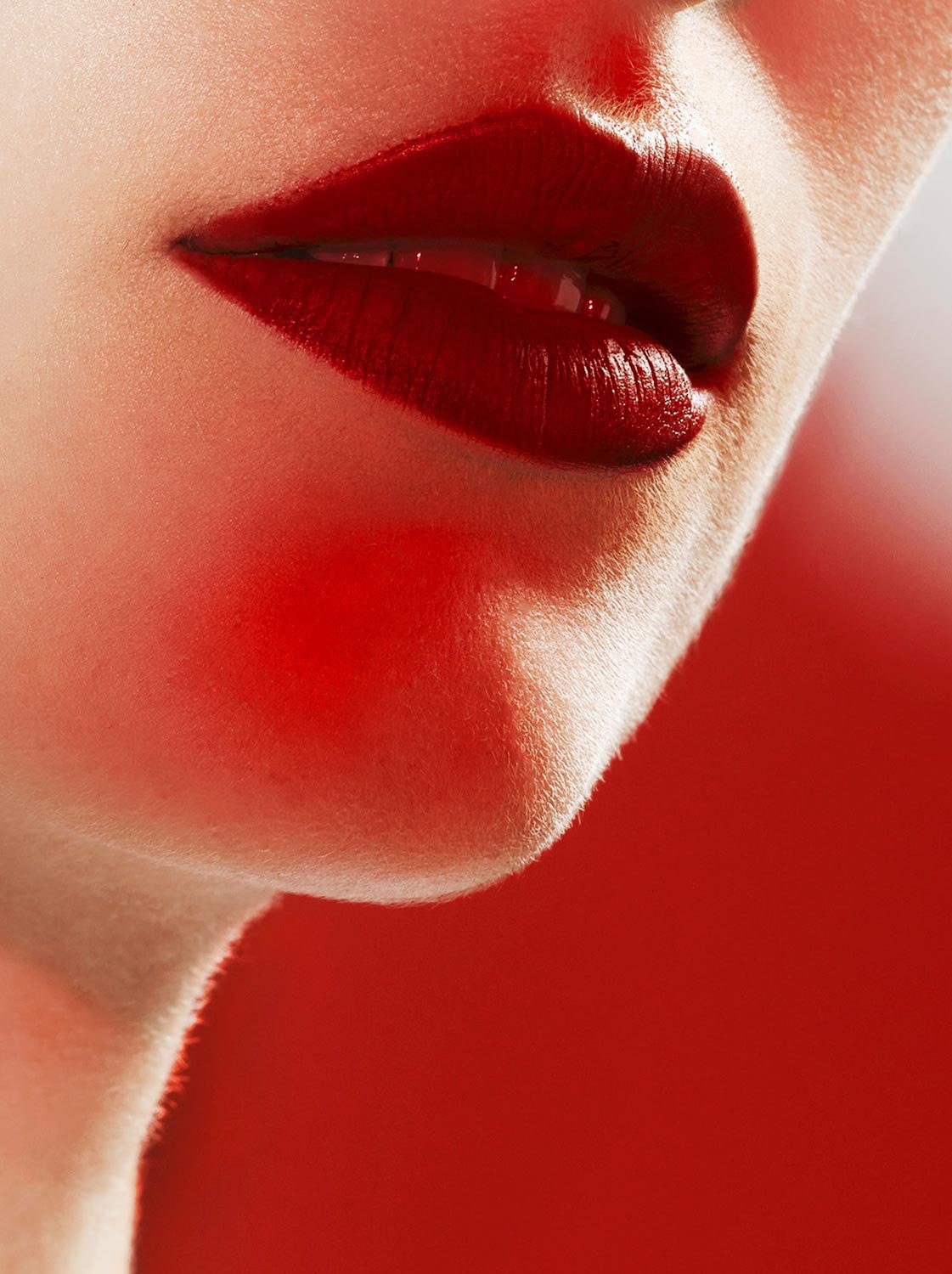 Close-up of a woman's face focusing on lips, with glossy red lipstick. The skin appears smooth with soft lighting and a blurred red background.