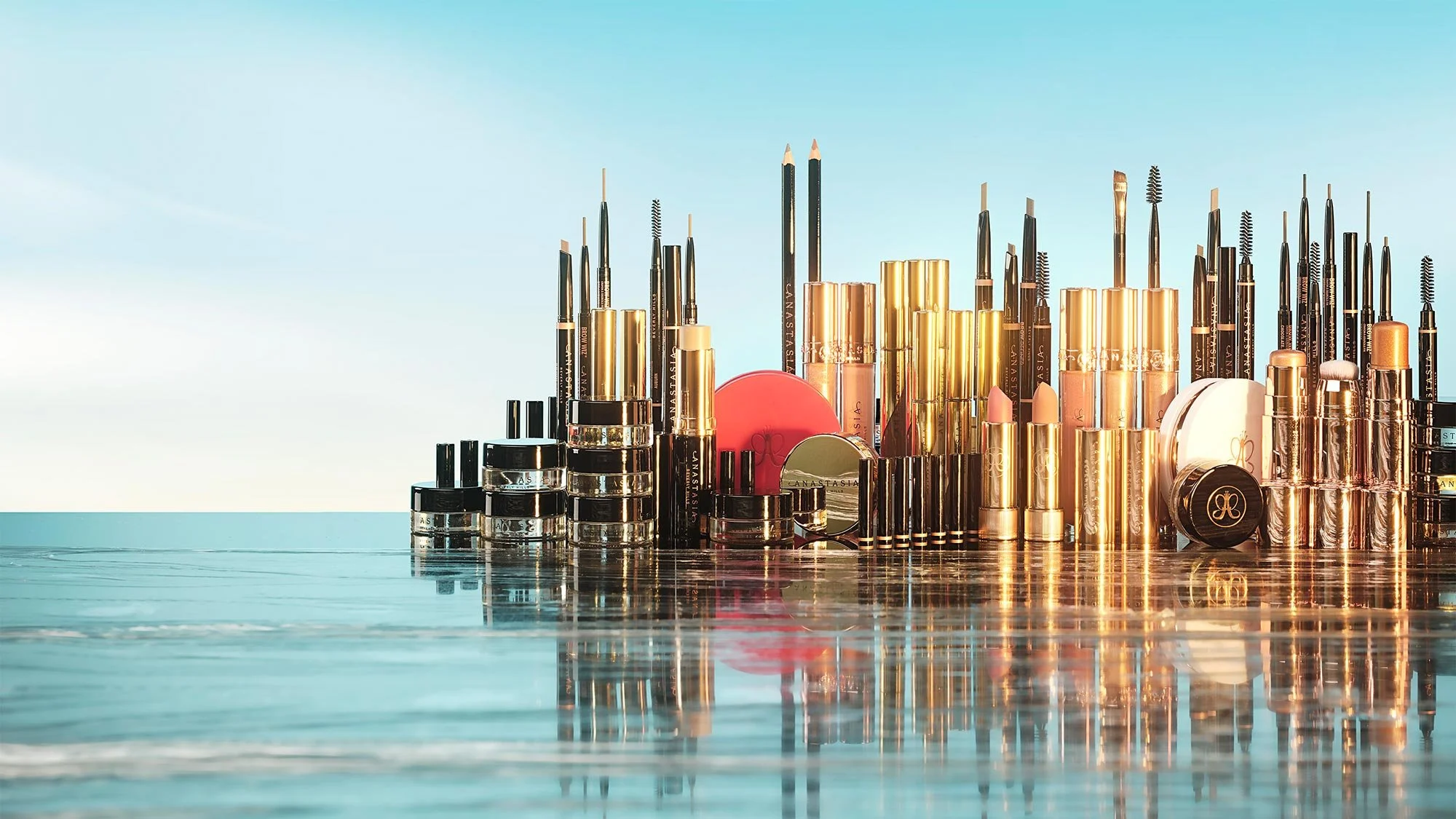 Assorted makeup products including eyeliners, lipsticks, and creams arranged on a reflective surface outdoors with clear sky background.
