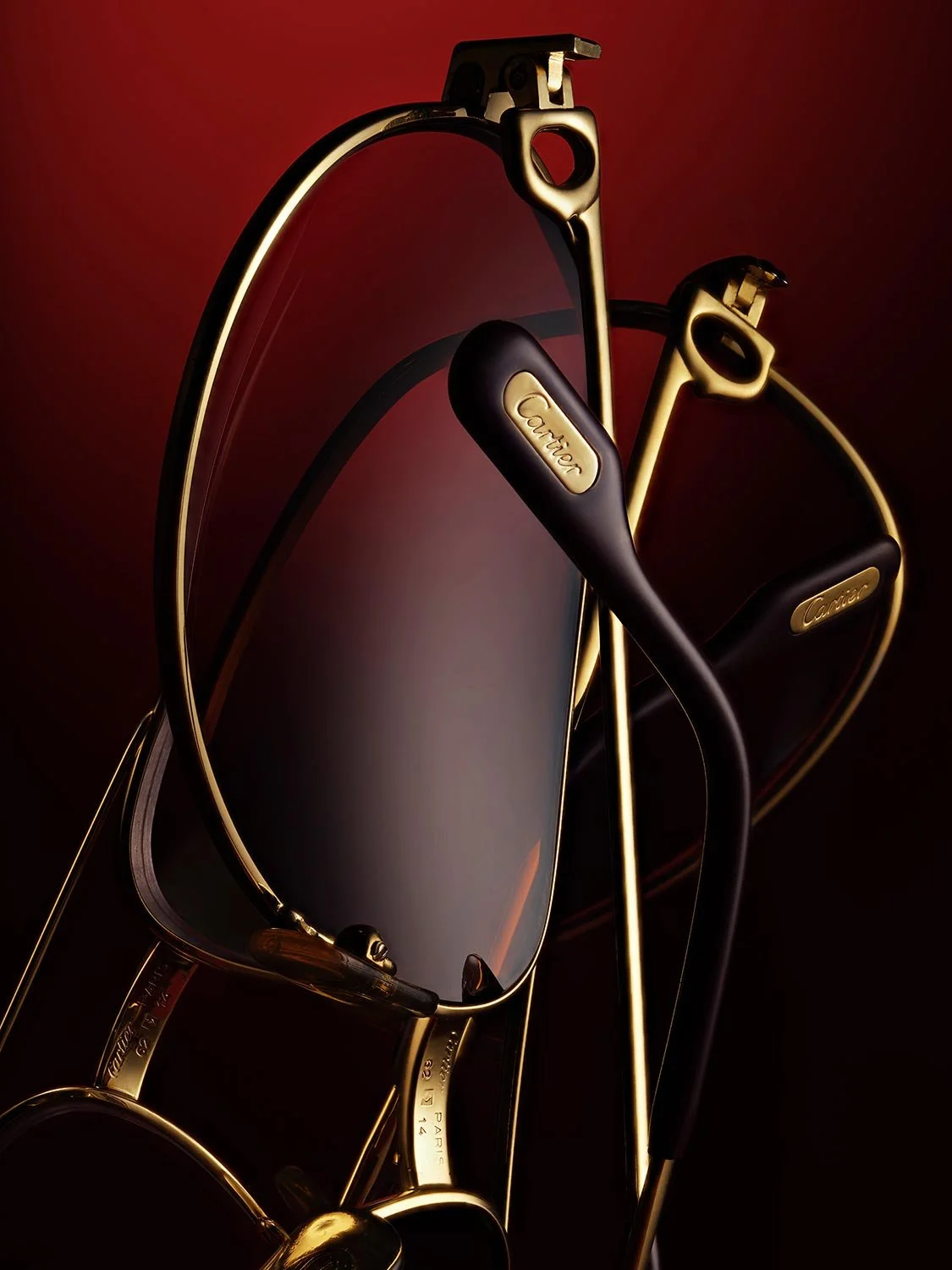Gold and black eyeglasses with a Cartier logo on the temples, reflective tinted lenses, set against a dark gradient red and black background.